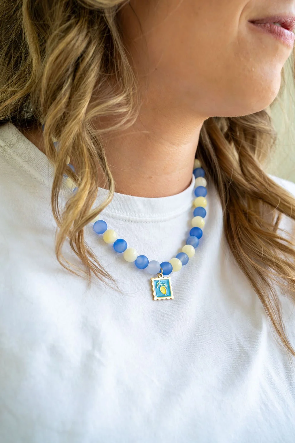 Close-up of a woman wearing a white shirt and a colorful beaded necklace with a square pendant featuring a lemon design.