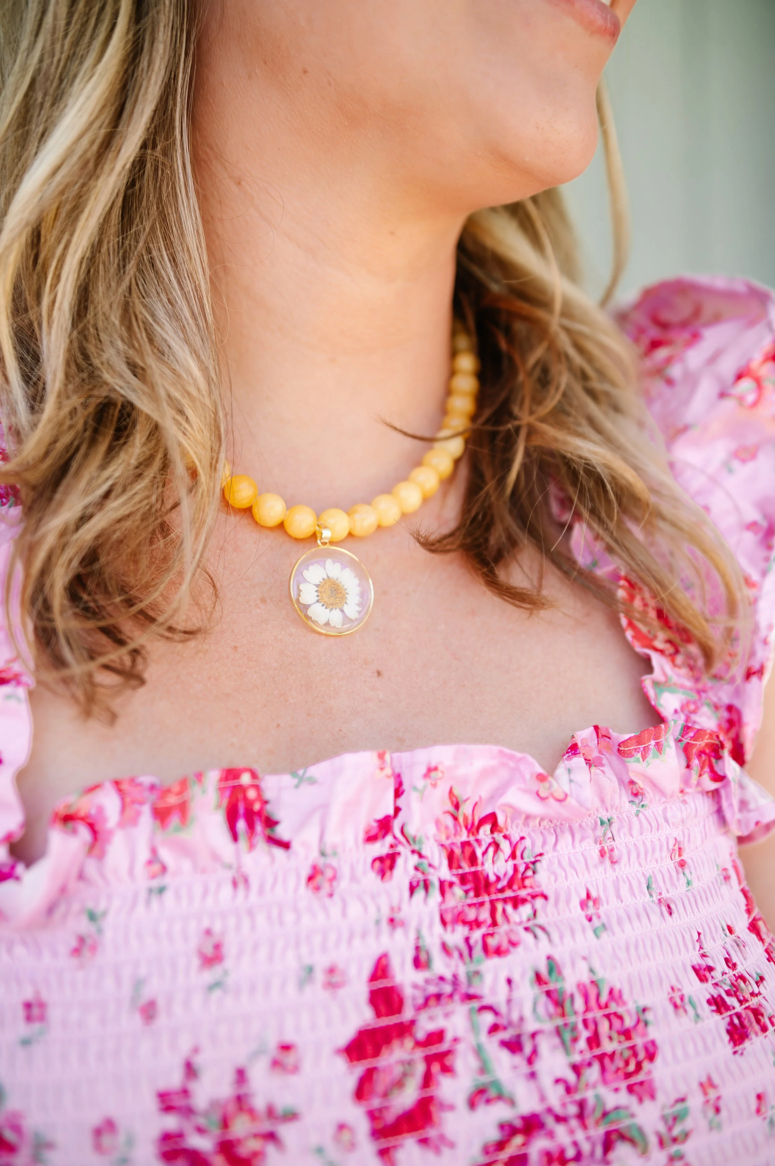 A close-up of a woman wearing a pink floral dress and a yellow beaded necklace with a daisy pendant.
