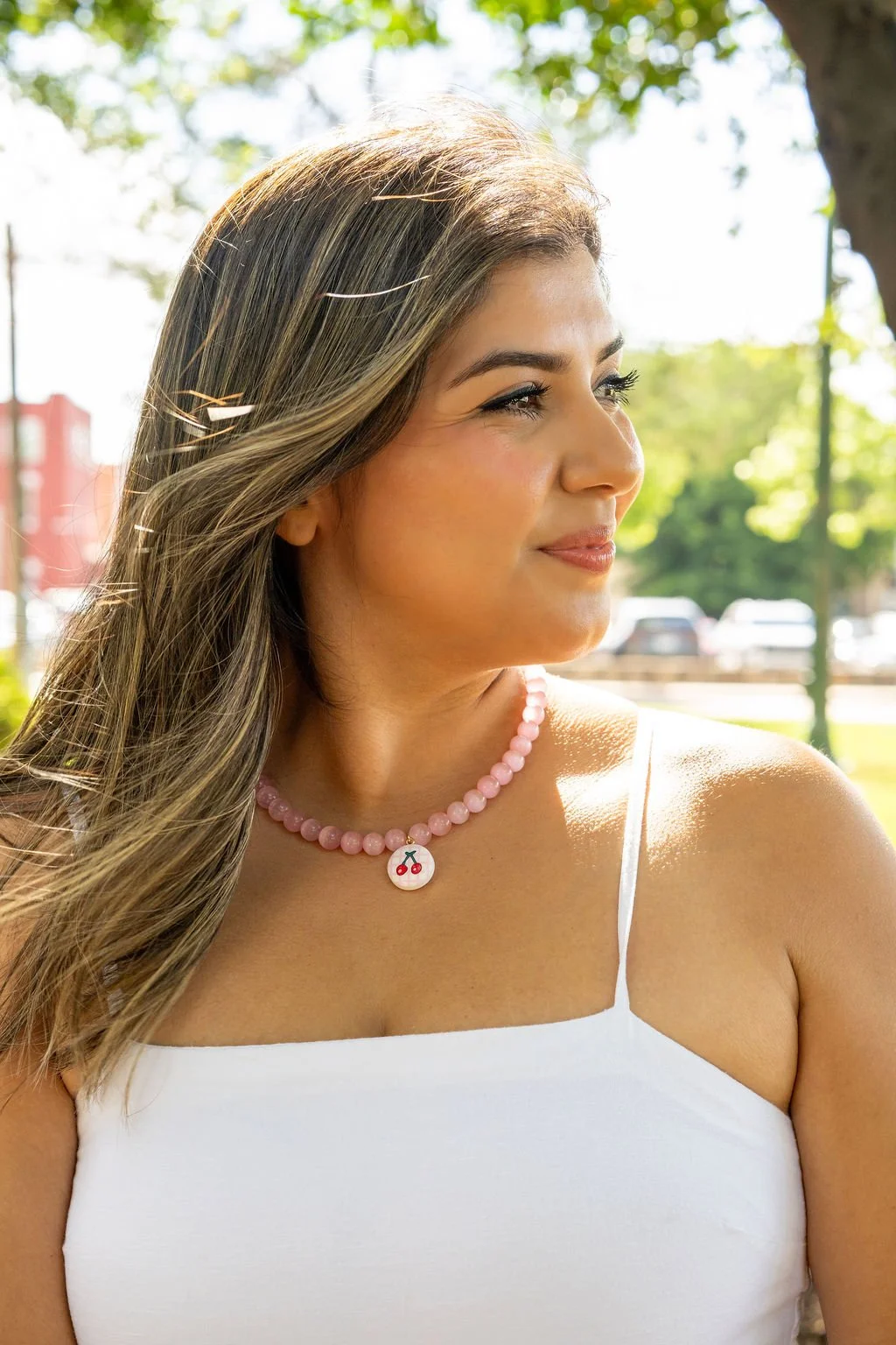 A woman with long wavy hair and a pink cherry necklace, wearing a white tank top, standing outdoors in a sunny park with trees and parked cars in the background.