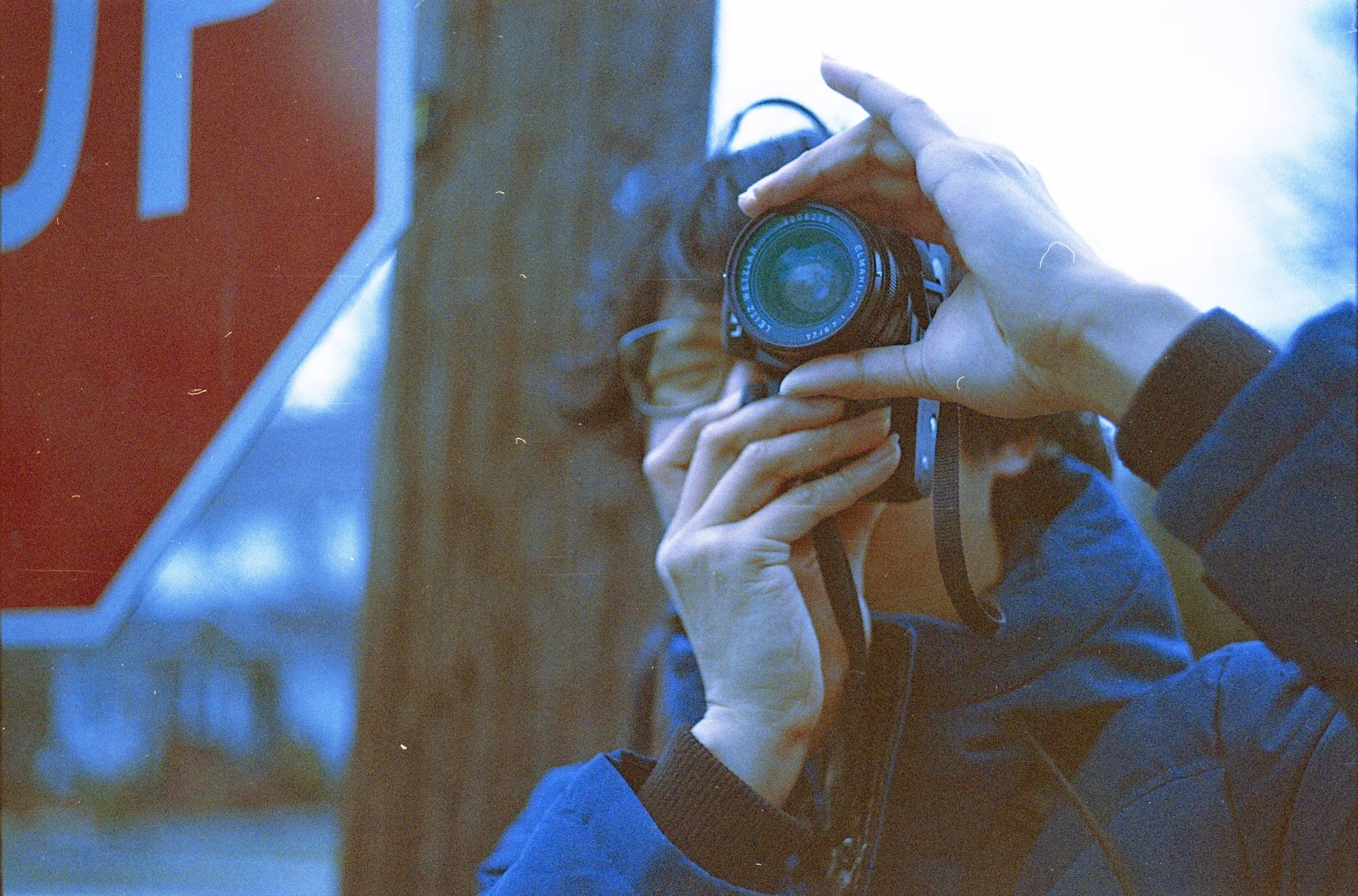 Person taking a photo in a mirror, showing their hand holding a camera, with a red sign and part of a glass window or display in the background.