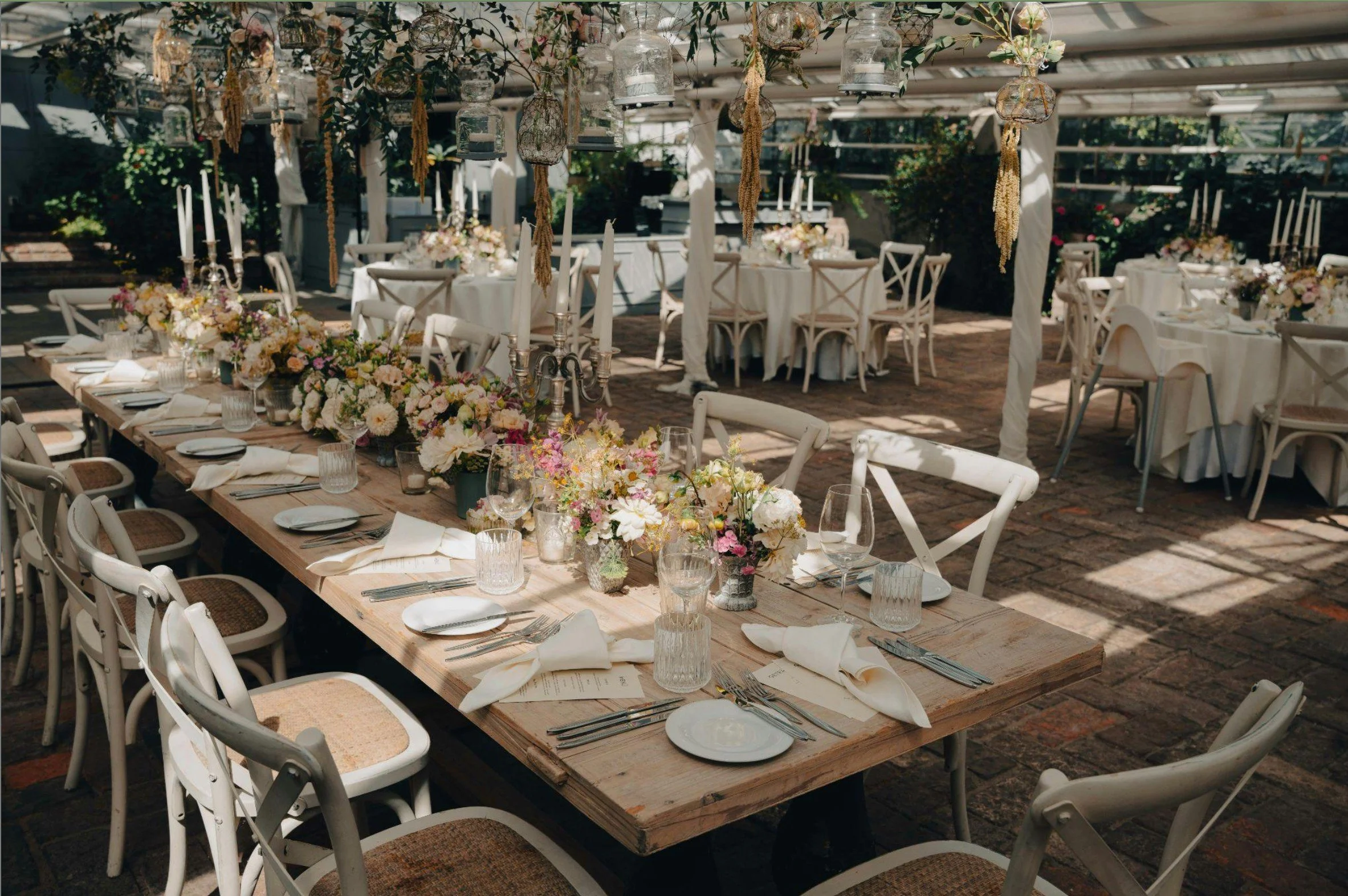 A beautifully decorated outdoor dining area with long wooden tables, white chairs, floral centerpieces, and hanging glass lanterns and flowers under a shaded patio.
