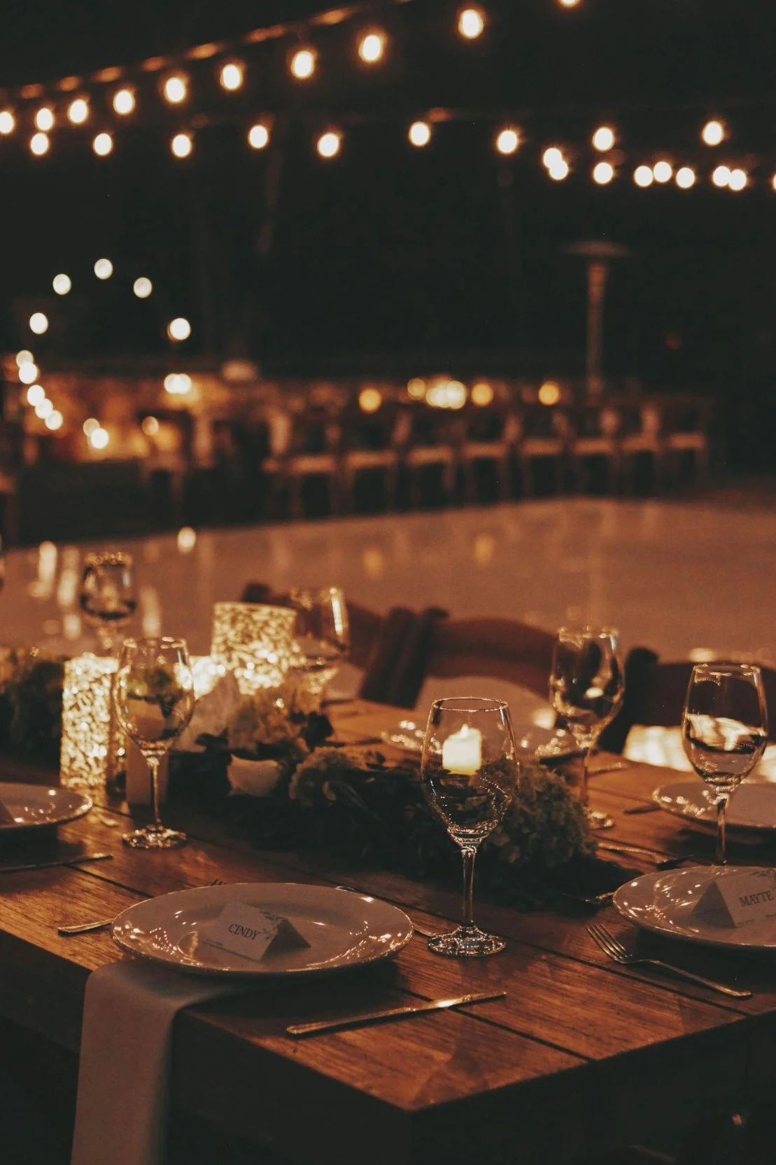 A dimly lit dinner table set for a special occasion with wine glasses, plates with name cards, and candle lanterns, in a decorated outdoor setting at night.