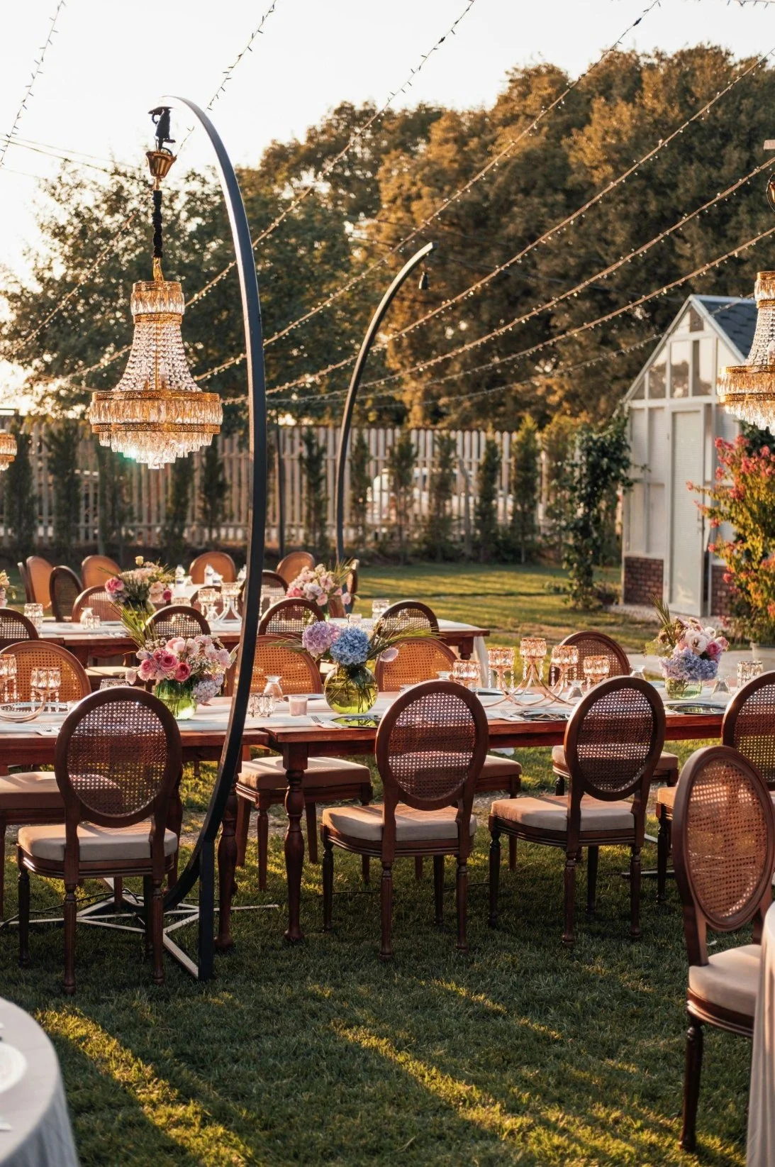 Outdoor wedding reception setup with long tables decorated with floral centerpieces, surrounded by wooden chairs, illuminated chandeliers, string lights, and a garden setting with trees and a white shed in the background.
