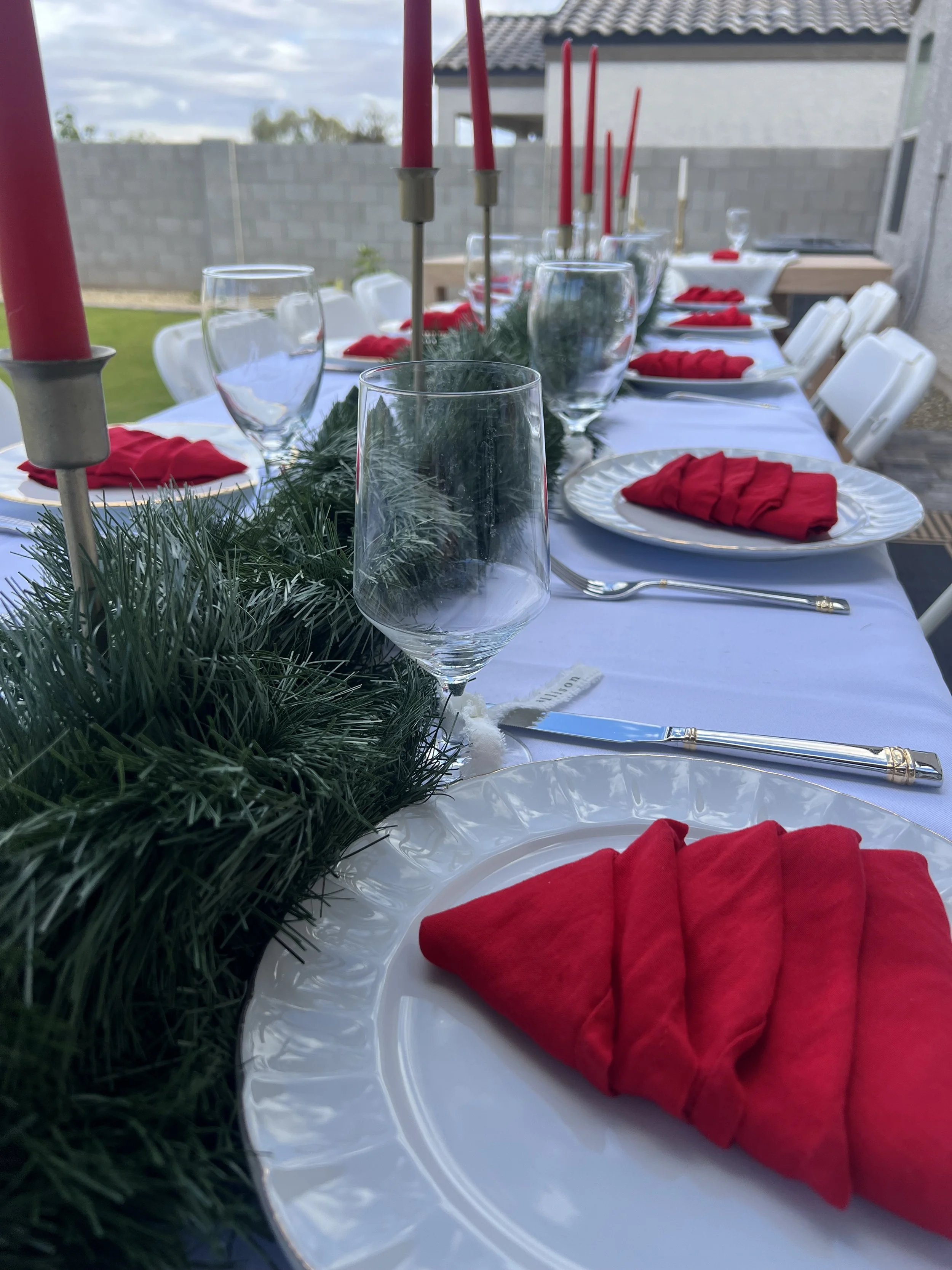 A long outdoor table set for a holiday gathering with white tablecloth, red napkins, glassware, candlesticks with red candles, and greenery centerpiece, with chairs arranged around the table.
