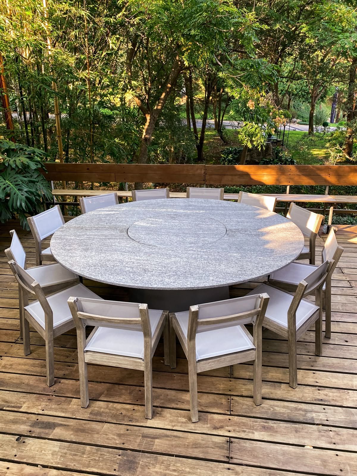 A round outdoor table with white and wood chairs on a wooden deck, surrounded by green trees and plants.