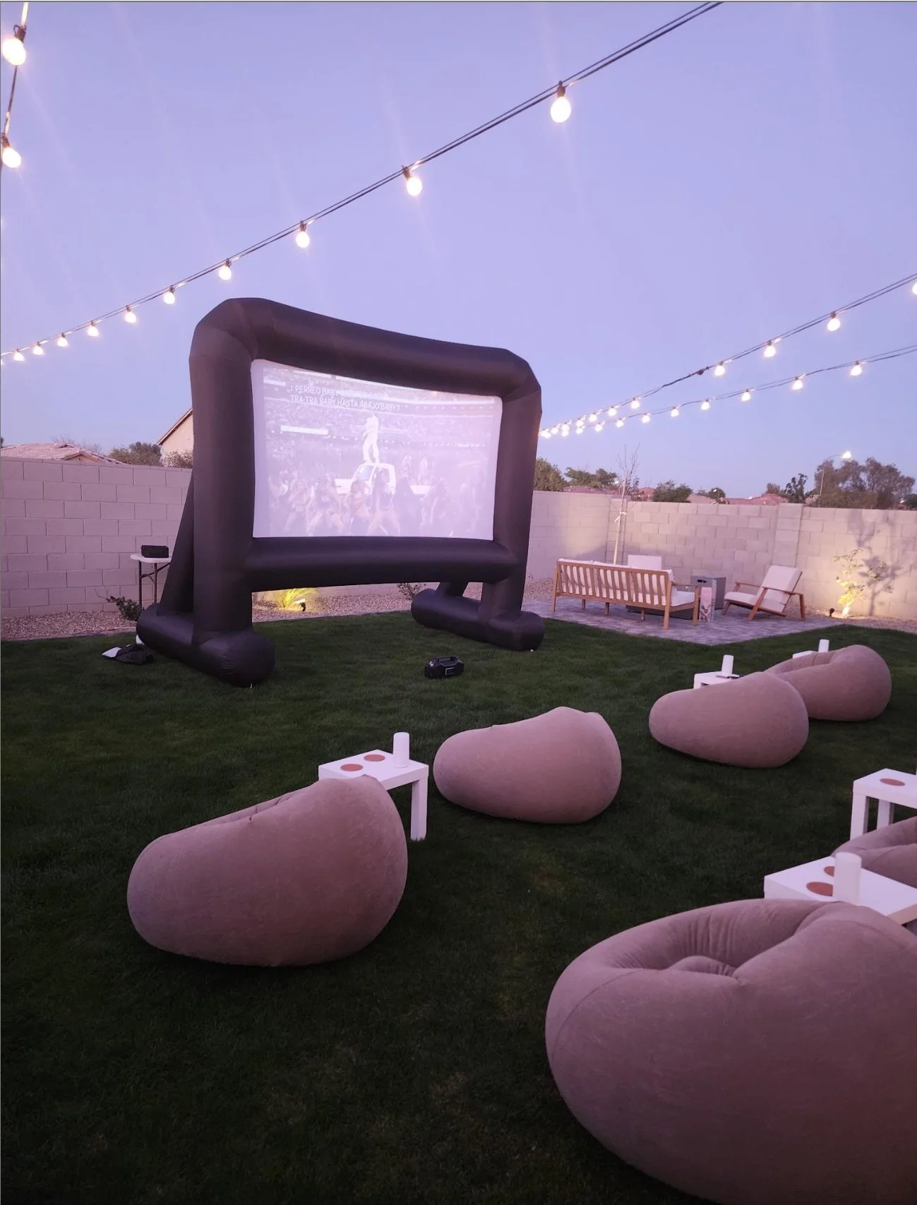 Outdoor backyard movie setup with a large inflatable screen, comfortable pink bean bag chairs, small white side tables with drinks, a cushioned sofa, and a couple of lounge chairs, illuminated by string lights overhead.
