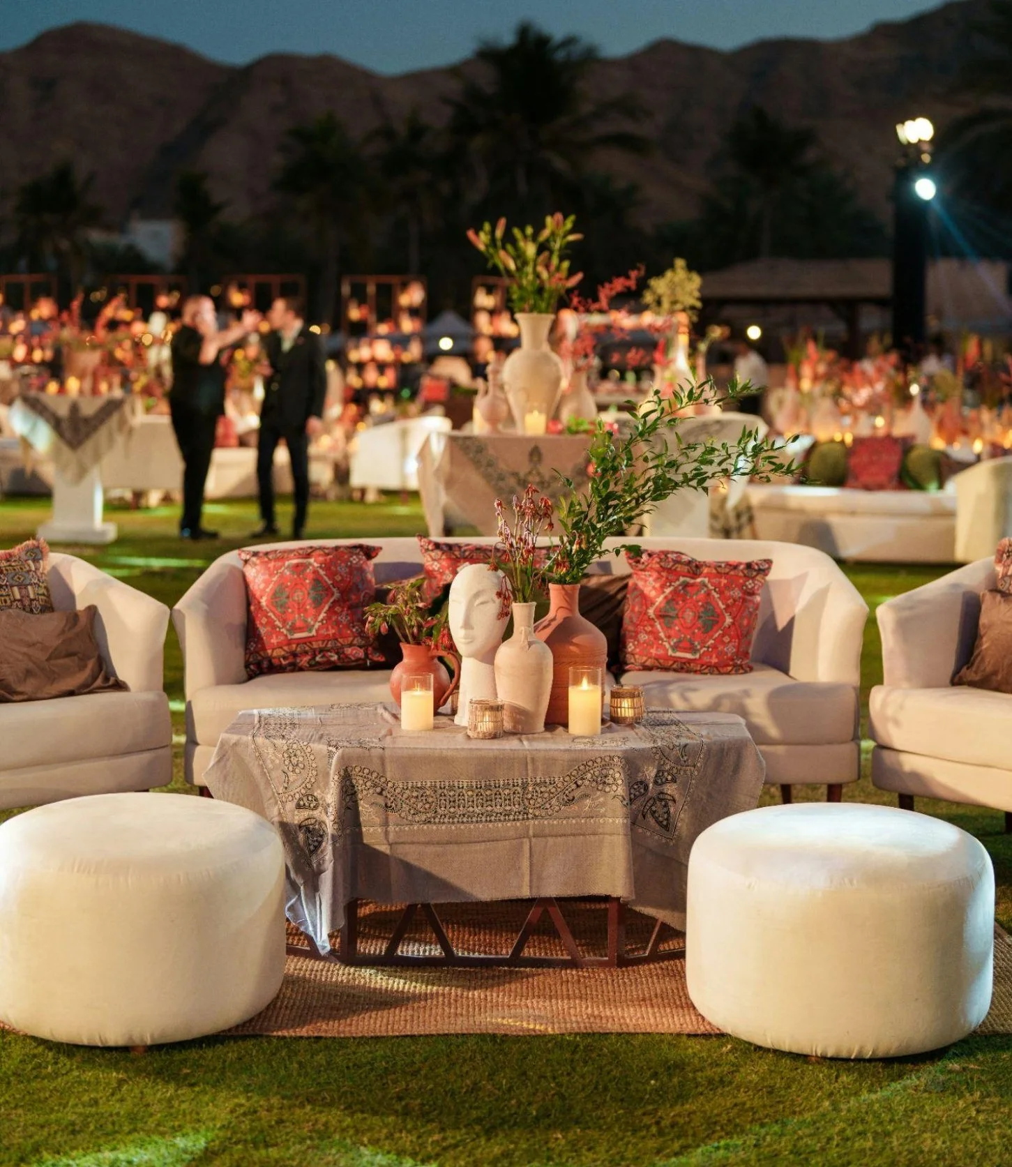 Outdoor evening event with elegant seating arrangements, decorated with candles, vases of flowers, and cushions, set against a backdrop of mountains and trees.