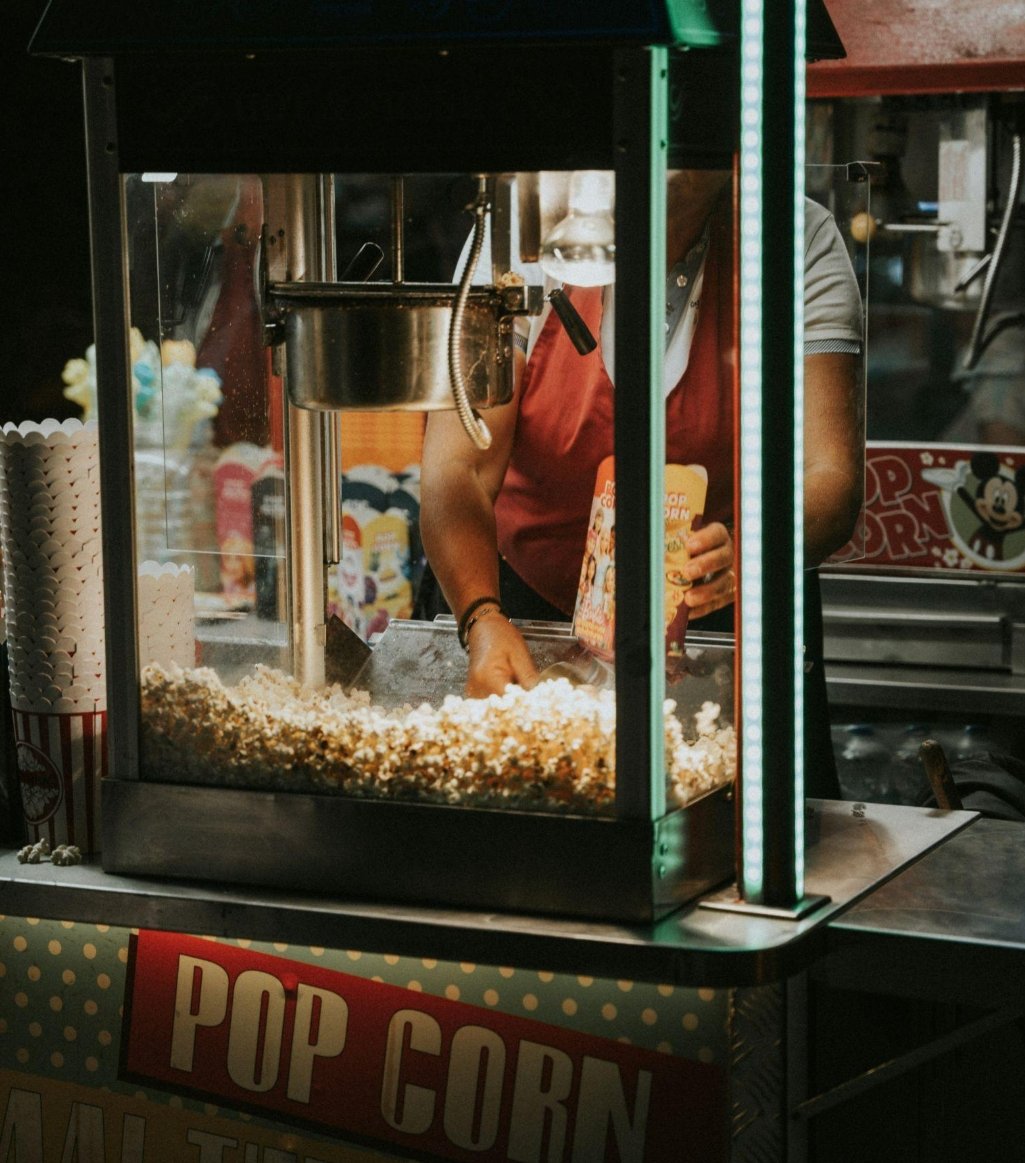 A popcorn stand at a fair or carnival with a person serving popcorn behind the glass. The person is holding a popcorn box, and popcorn is visible inside the stand.