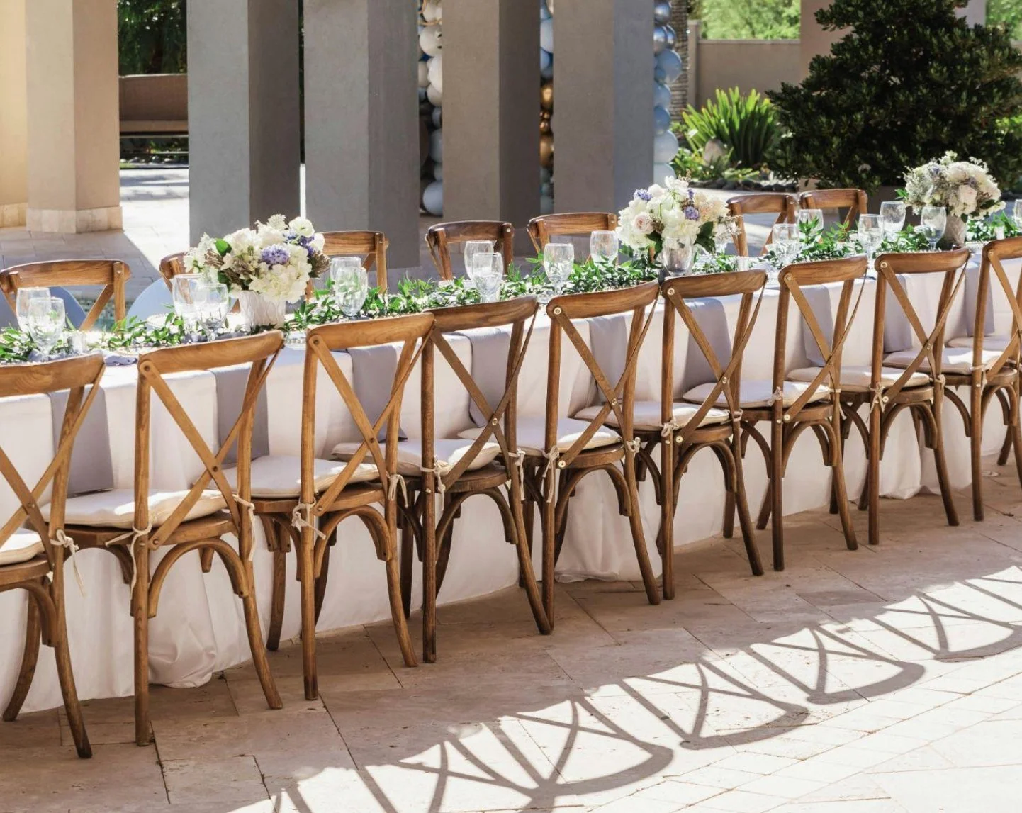 Long rectangular outdoor dining table set with white tablecloths, wooden cross-back chairs with white cushions, flower centerpieces, and glassware, in a garden setting with trees and greenery.