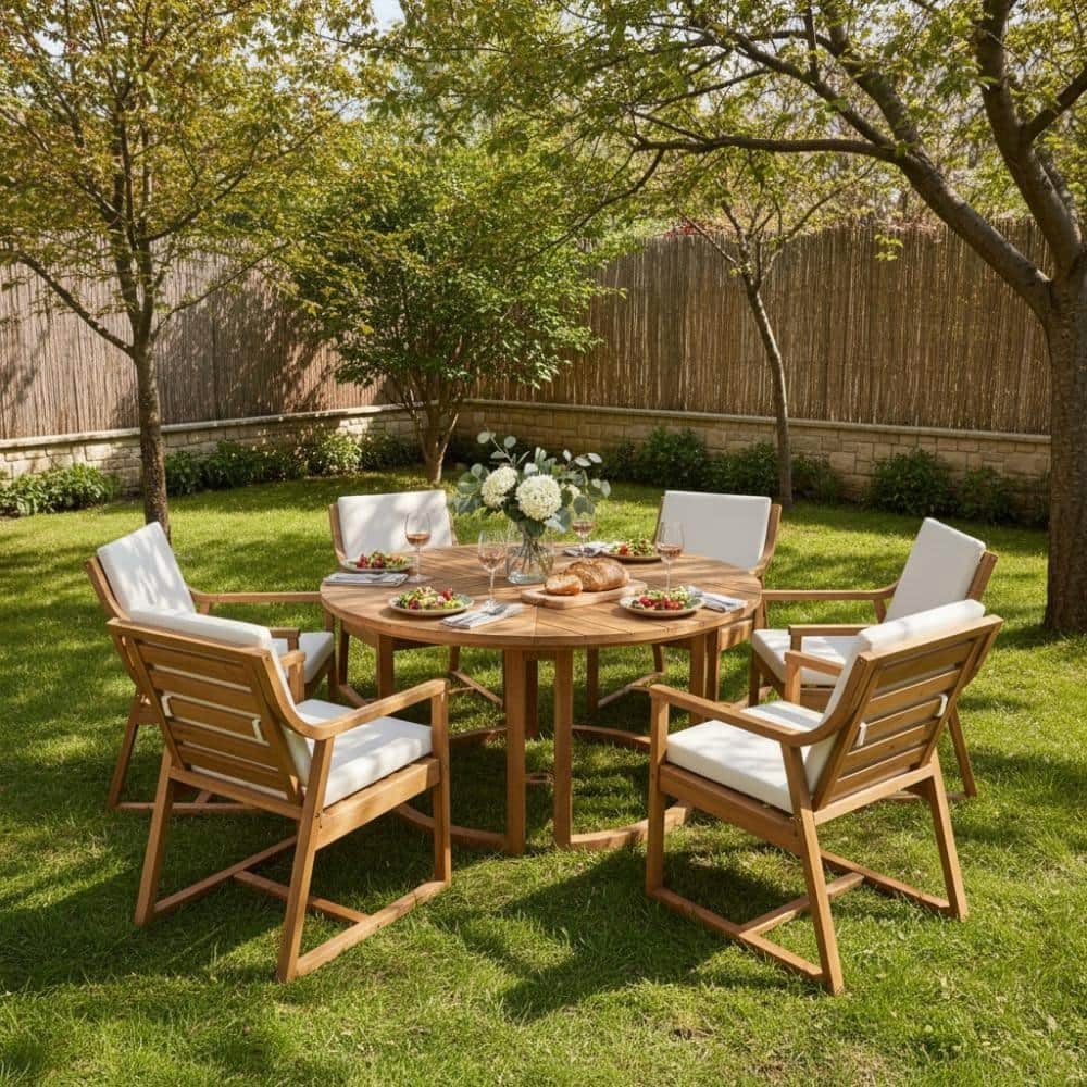 Outdoor garden scene with a round wooden table set for a meal, surrounded by six wooden chairs with white cushions. The table has a vase of white flowers, wine glasses, plates of food, bread, and napkins. There are trees and a wooden fence in the background on a sunny day.