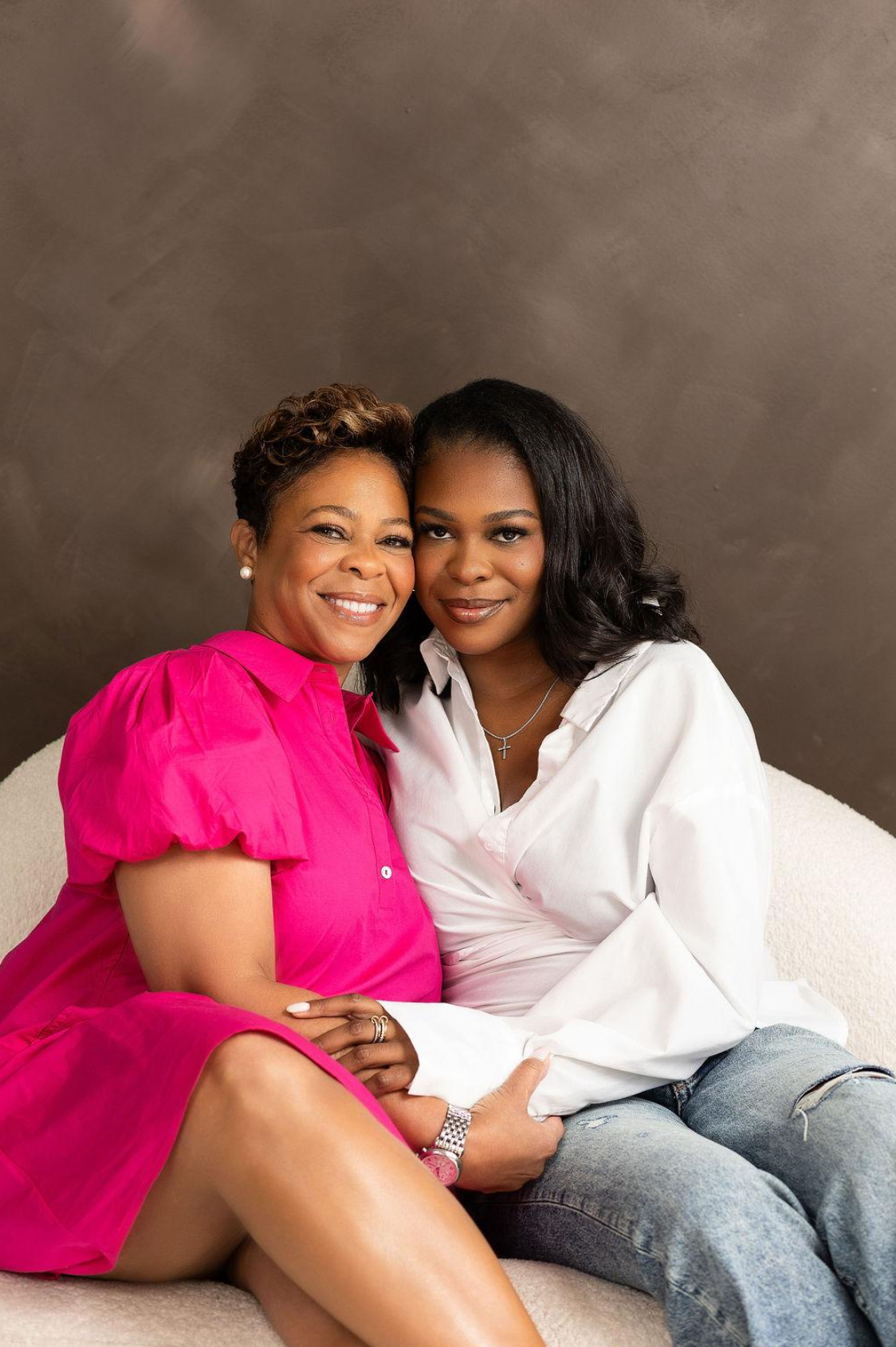 Two women sitting close together on a beige couch, smiling at the camera, one wearing a pink dress and the other in a white shirt and jeans, with a dark brown background.