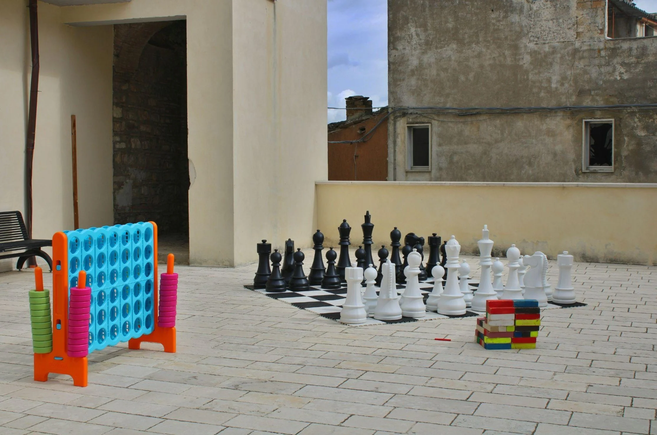 Outdoor courtyard with large chess and checkers game set on tiled patio, colorfulConnect Four game, and a stack of small wooden blocks, with old buildings in the background and cloudy sky.