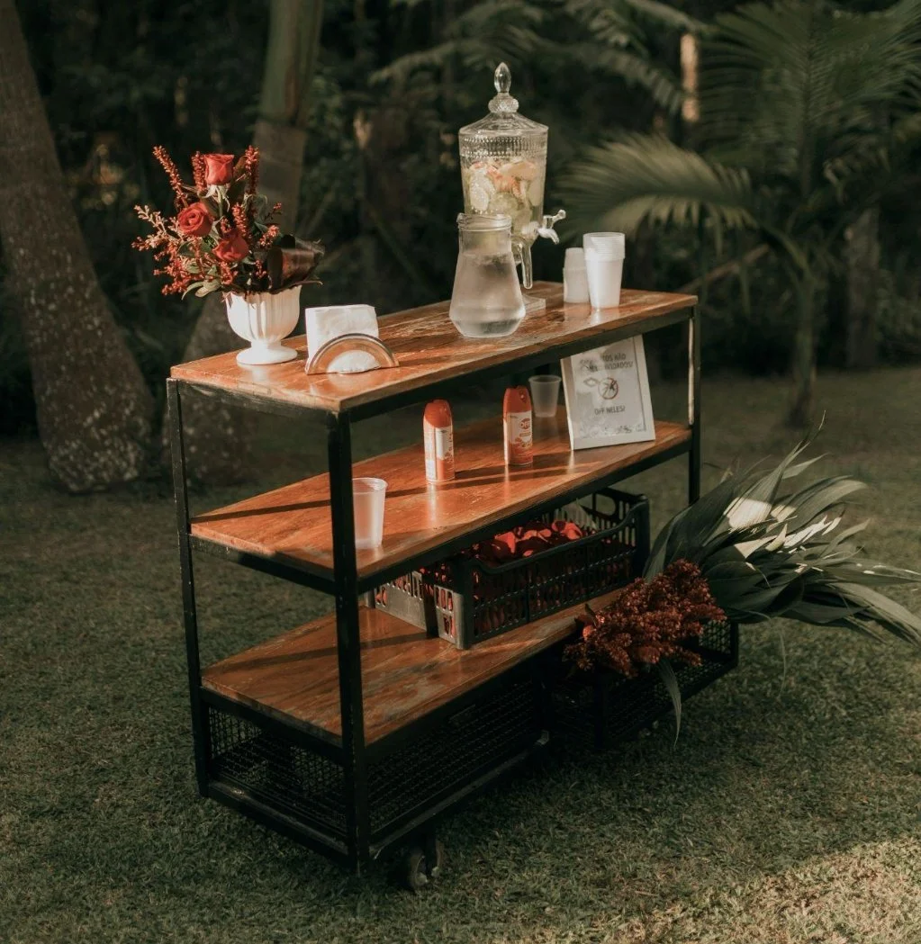 A wooden cart with three shelves is set on grass in a garden with trees and plants in the background. The top shelf has a floral arrangement, glass jar with a spigot filled with a citrus drink, cups, and a card. The middle shelf has two bottles and a framed sign. The bottom shelf has a black basket with red items and some large green leaves and flowers on the ground beside the cart.