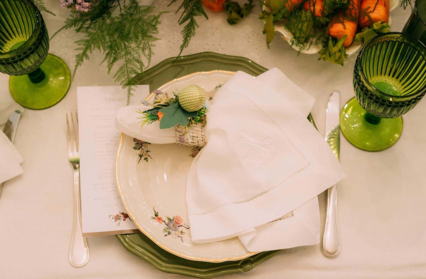 Elegant table setting with a green plate, white cloth napkin, and a small decorative basket with a green checkered ball. Green glasses and silverware are arranged around the plate, with a menu or program card underneath. Fresh greenery and a bowl of carrots are visible as part of the table decor.