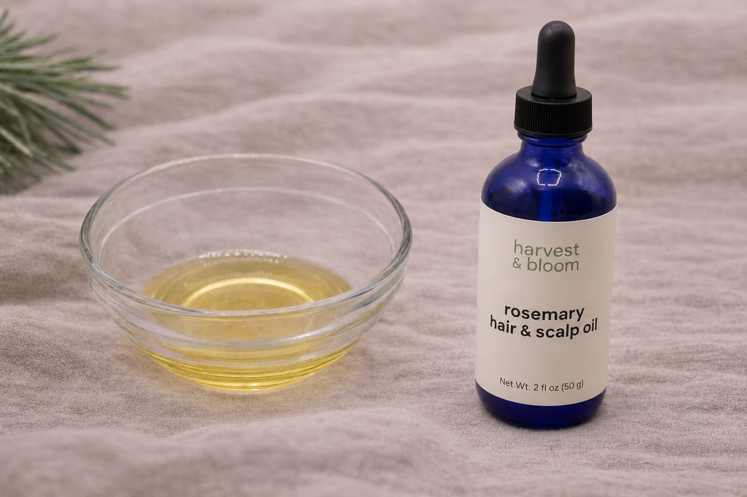 A close-up view of golden, slow-infused rosemary hair oil in a small glass bowl next to an individual cobalt blue glass dropper bottle of Harvest & Bloom scalp oil.