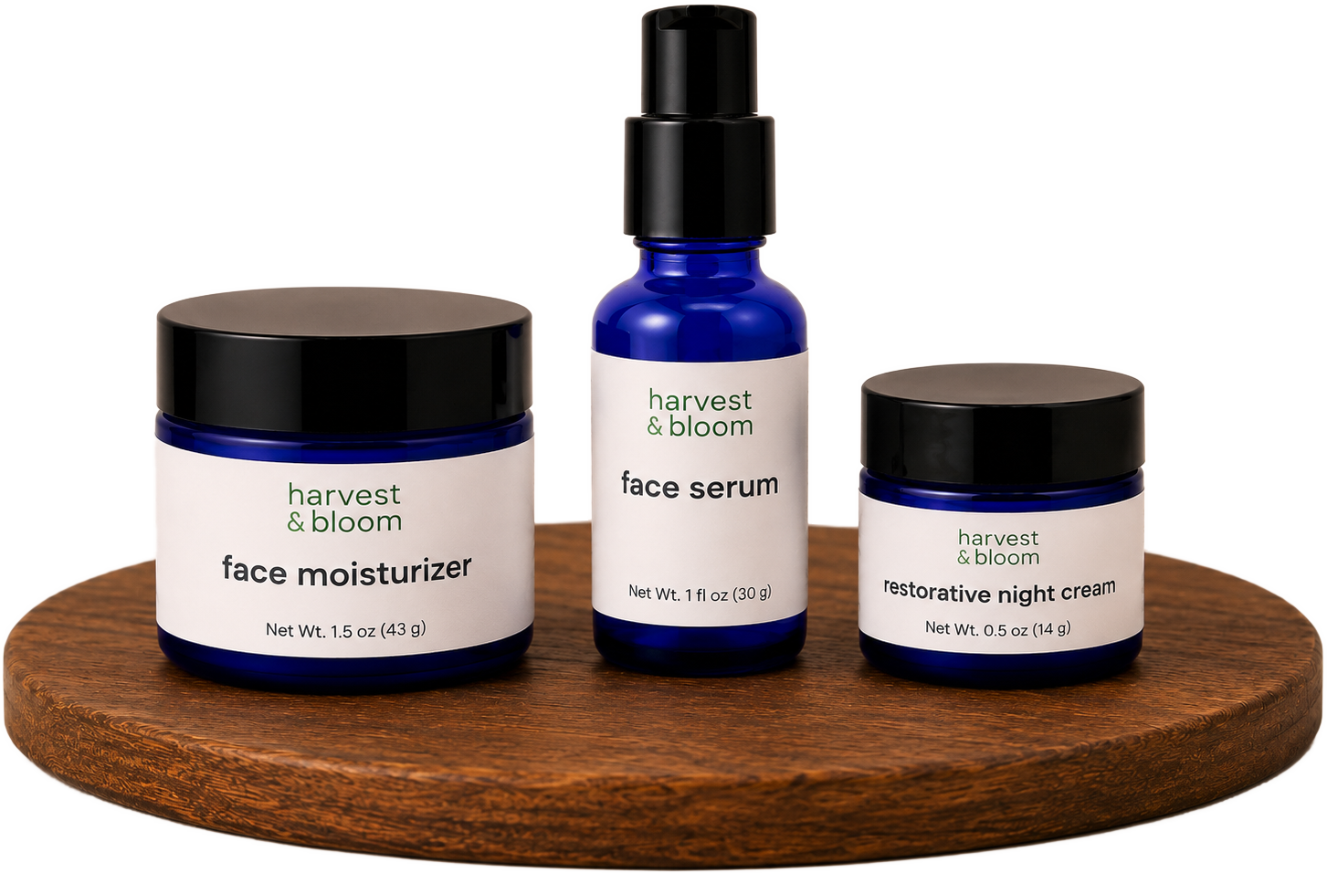Three cobalt blue glass containers of Harvest & Bloom waterless facial skincare products, including face moisturizer, face serum, and restorative night cream, displayed on a round wooden riser against a warm cream background.