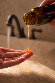 Golden raw honey liquid pouring from an amber glass dropper bottle into a wet open hand over a bathroom sink with a blurred running water faucet.