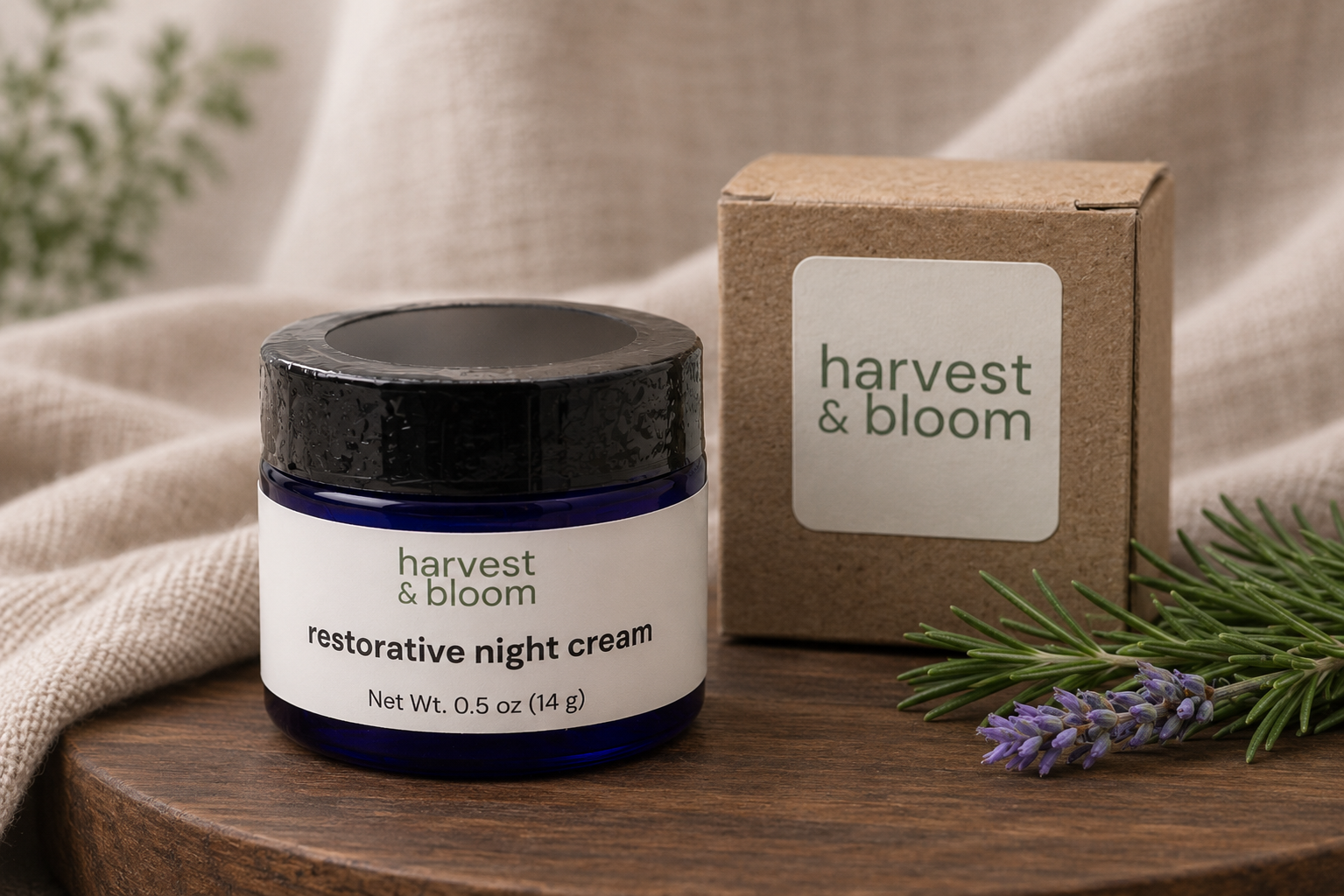 A front-facing view of a cobalt blue glass jar of Harvest & Bloom restorative night cream next to its minimalist kraft paper packaging box on a round wooden tray with fresh botanical sprigs.