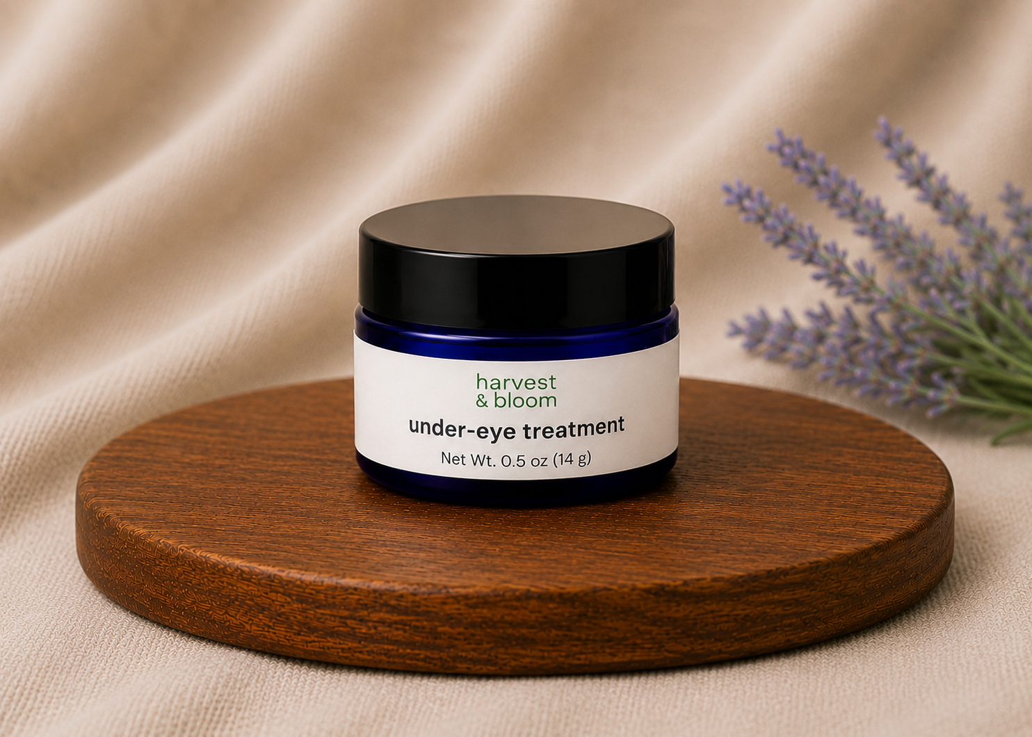 Harvest and Bloom natural under-eye treatment in a small blue glass jar with a black lid against a soft fabric backdrop.