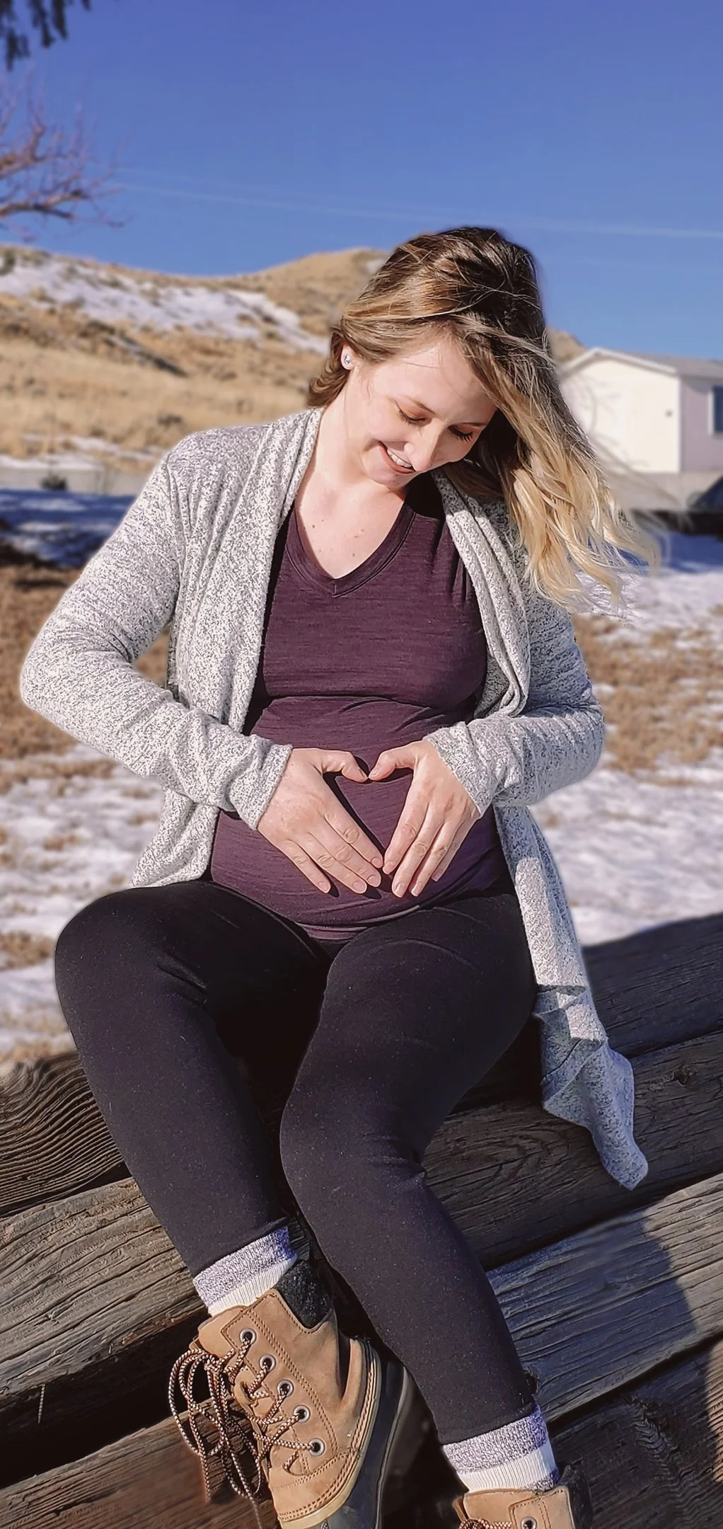 A woman with blonde hair sitting on a wooden log outdoors in winter, wearing a purple shirt, gray cardigan, black pants, tan boots, and forming a heart shape over her pregnant belly with her hands, smiling and looking down.