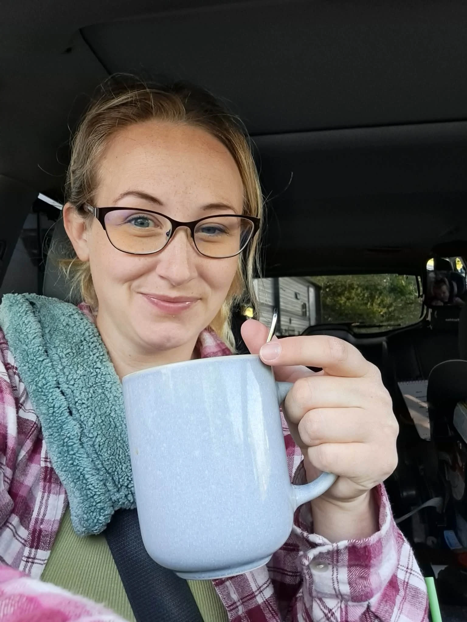 A woman with glasses smiling and holding a large white mug in a vehicle, with a seatbelt across her chest and blurred outdoor scenery visible through the window.