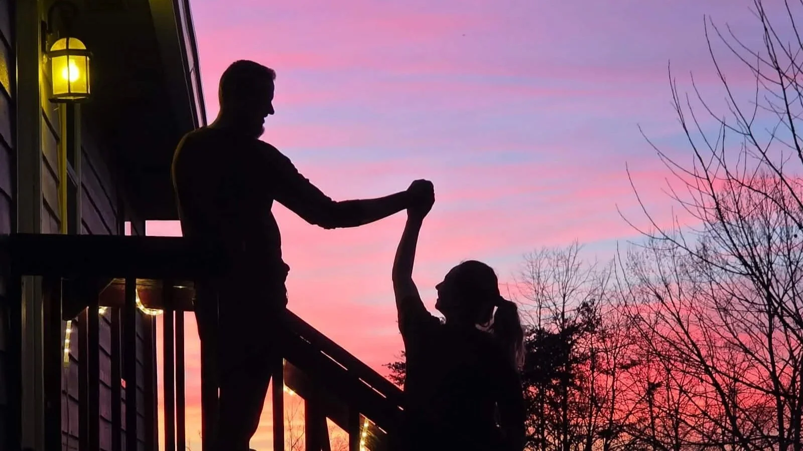 Silhouette of a man helping a woman up the stairs on a porch at sunset, with pink and purple sky and trees in the background.
