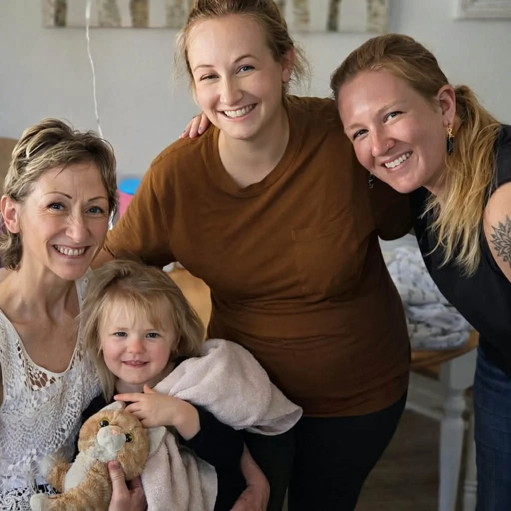 Four women, including a young girl holding a stuffed animal, are smiling and posing together in a cozy indoor setting.