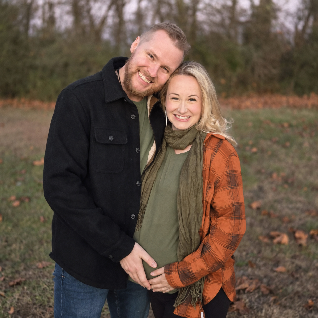 A smiling couple outdoors in autumn, with the man embracing the pregnant woman who is holding her belly, in a park with fallen leaves and trees in the background.
