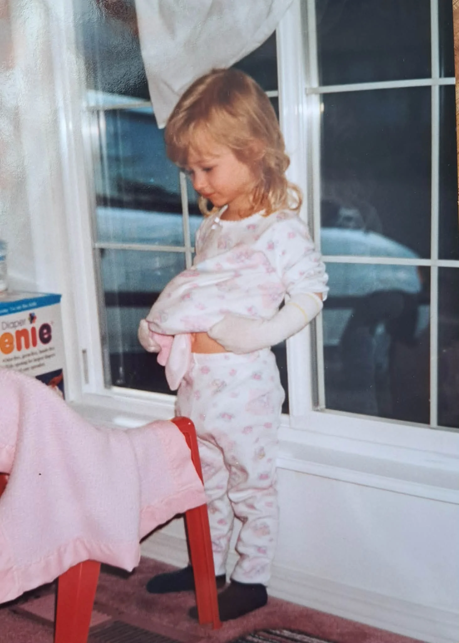 A young girl with red hair in pajamas with pink hearts, sitting in a room near a window, holding a small object, with a partial view of a person wearing a pink sleeve and red wristband holding her hand.