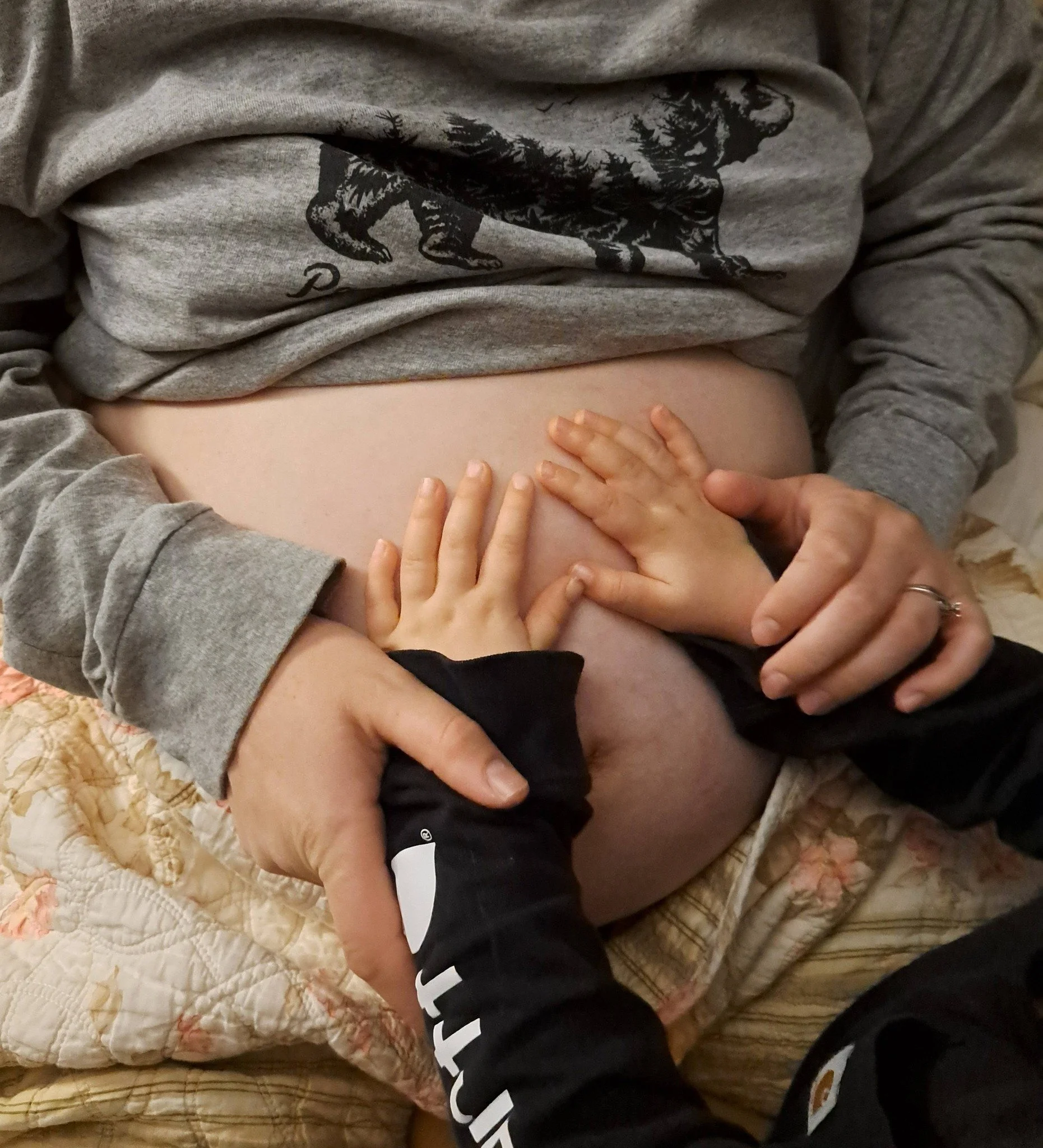 A person lying on a bed wearing a grey sweatshirt with a black graphic of a dog and a forest scene. Their hand is resting on their bare stomach, and another hand is touching the person's side. The bed has a quilted beige and pink floral blanket.