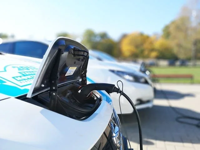 Close-up of an electric vehicle charging port with a plugged-in charger, cars in the background, with trees and a park.