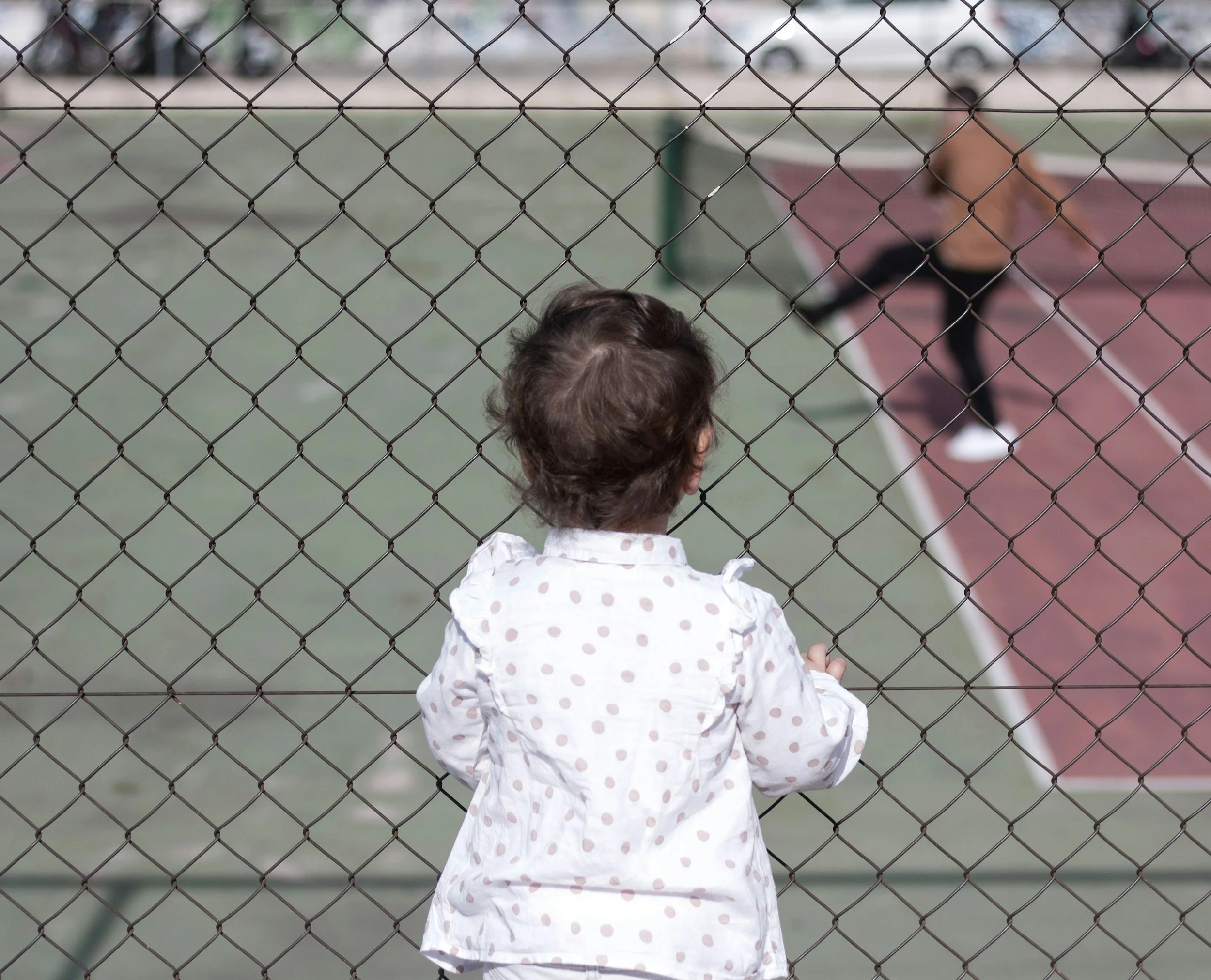 A young girl with curly hair in a white polka dot jacket stands behind a chain-link fence at a tennis court, watching a woman playing tennis in the background.