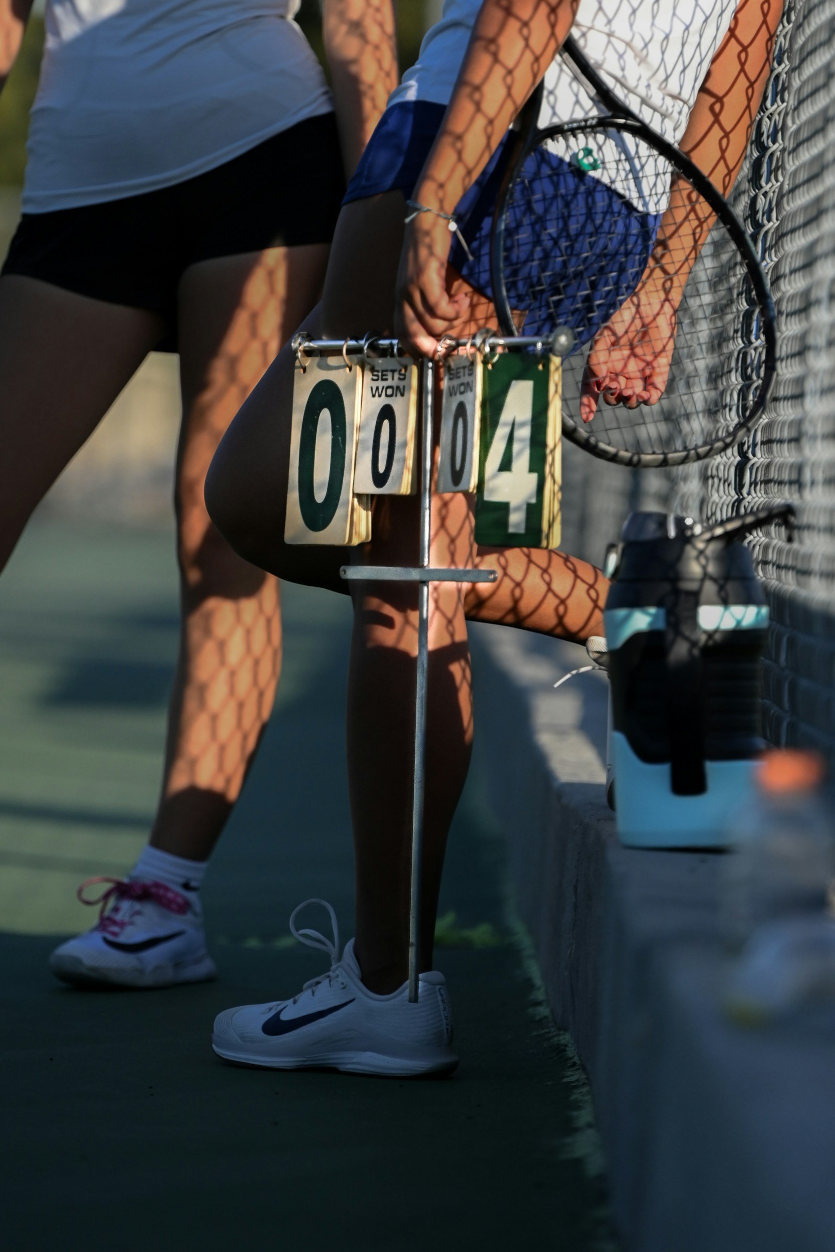 Close-up of a tennis score board displaying '00-04' on a tennis court. Two players in athletic gear, one with a tennis racket, are seen from the waist down, with their shadows cast on the ground and chain-link fencing in the background.