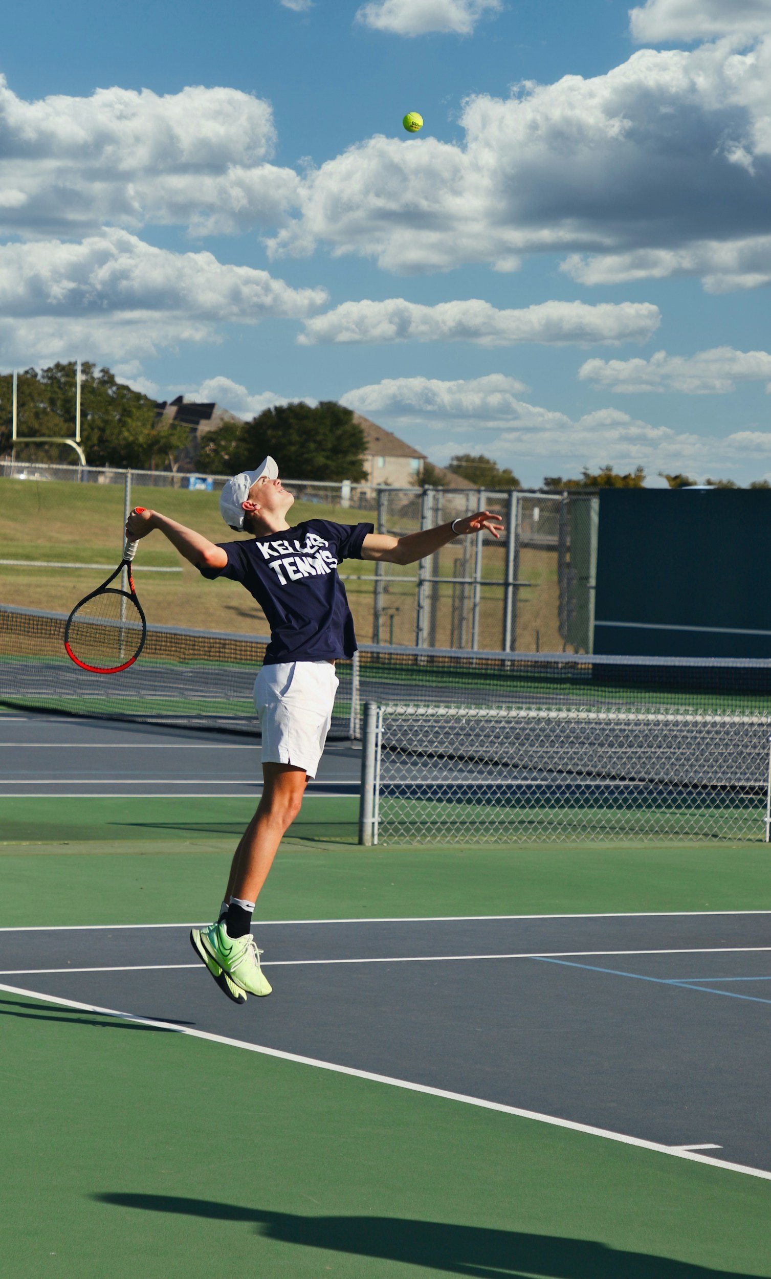 A young boy in tennis attire jumping to hit a tennis ball on an outdoor tennis court under a partly cloudy sky.