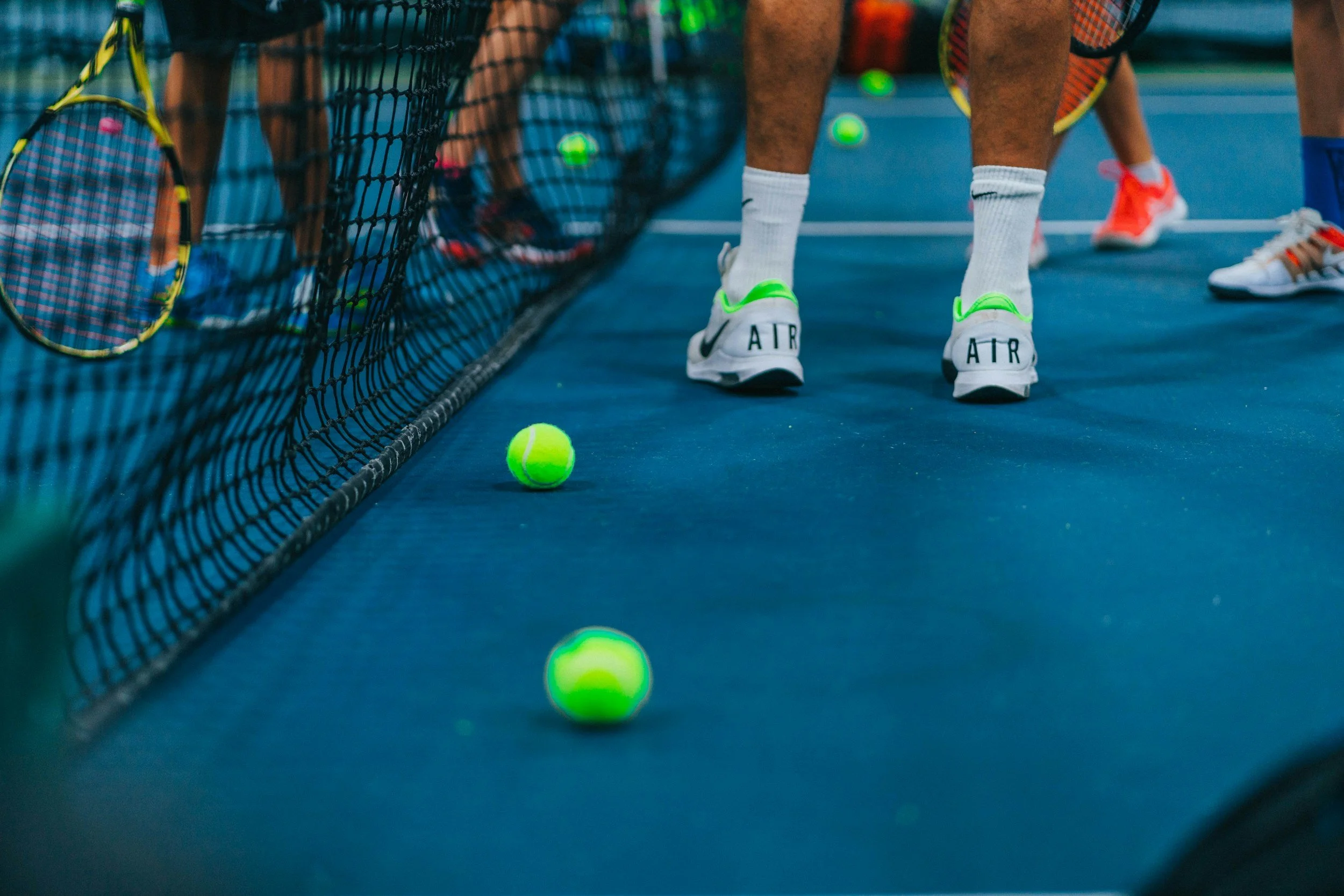 Multiple tennis balls scattered on a blue tennis court, with tennis rackets leaning against a black net and players standing nearby.