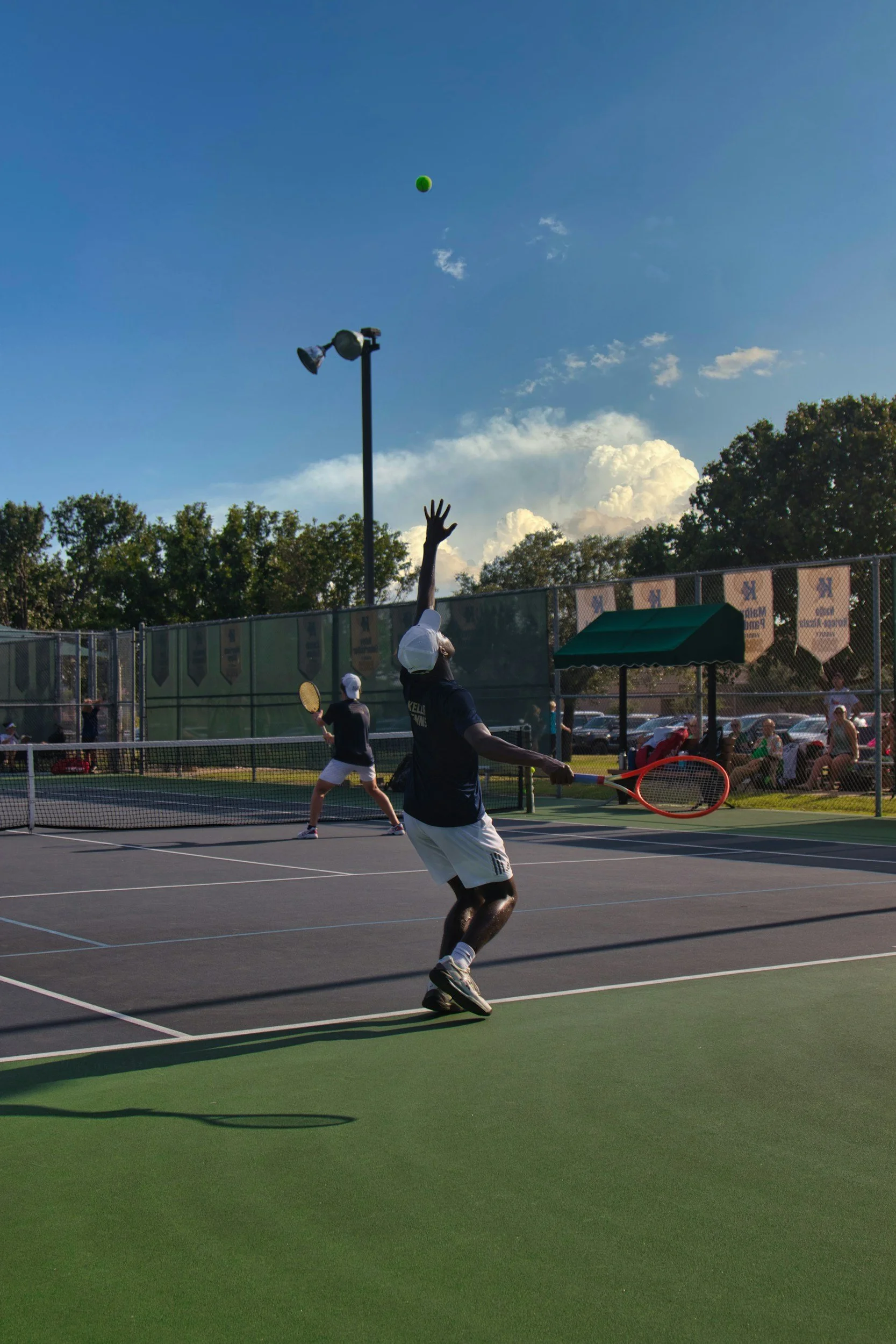 Tennis players engaging in a doubles match on an outdoor tennis court during the daytime, with one player reaching to hit the tennis ball that is in midair, surrounded by spectators and a clear sky with some clouds.
