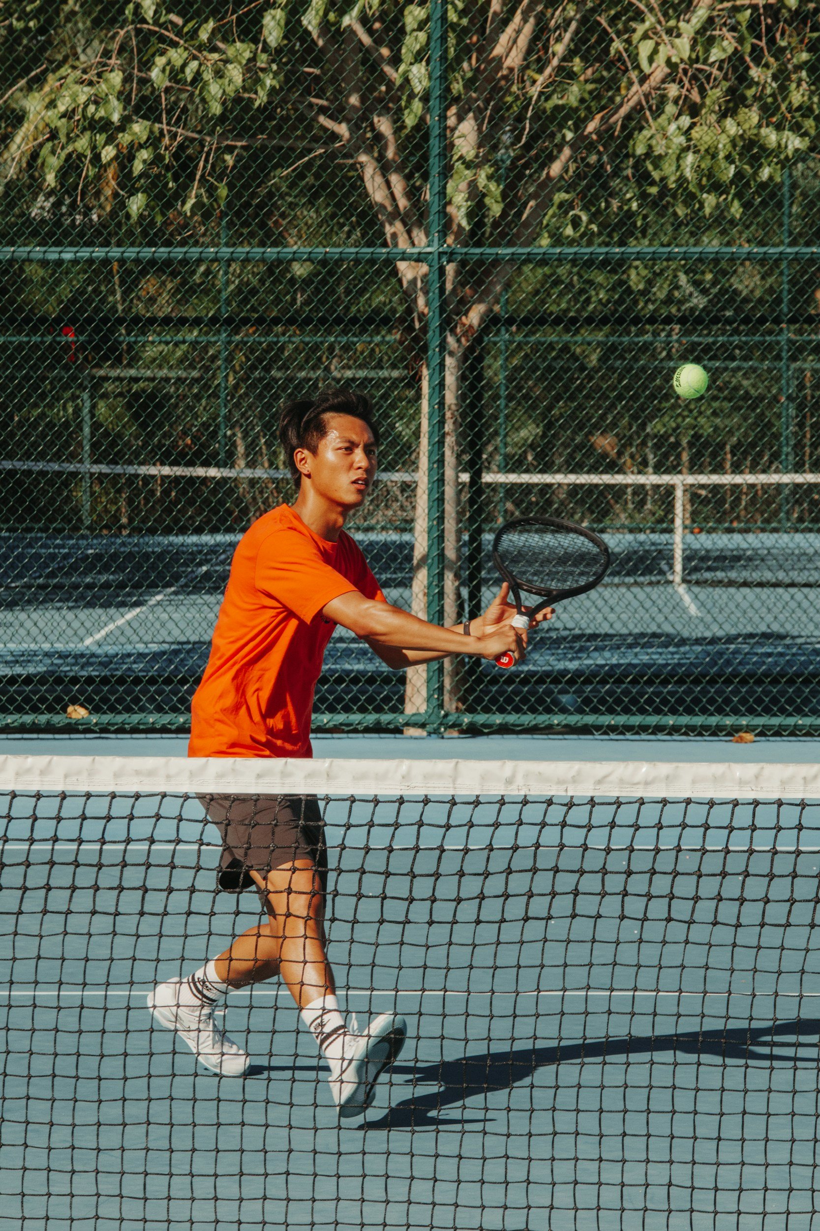 A young man in an orange shirt and black shorts playing tennis on an outdoor court, hitting a ball with a racket.