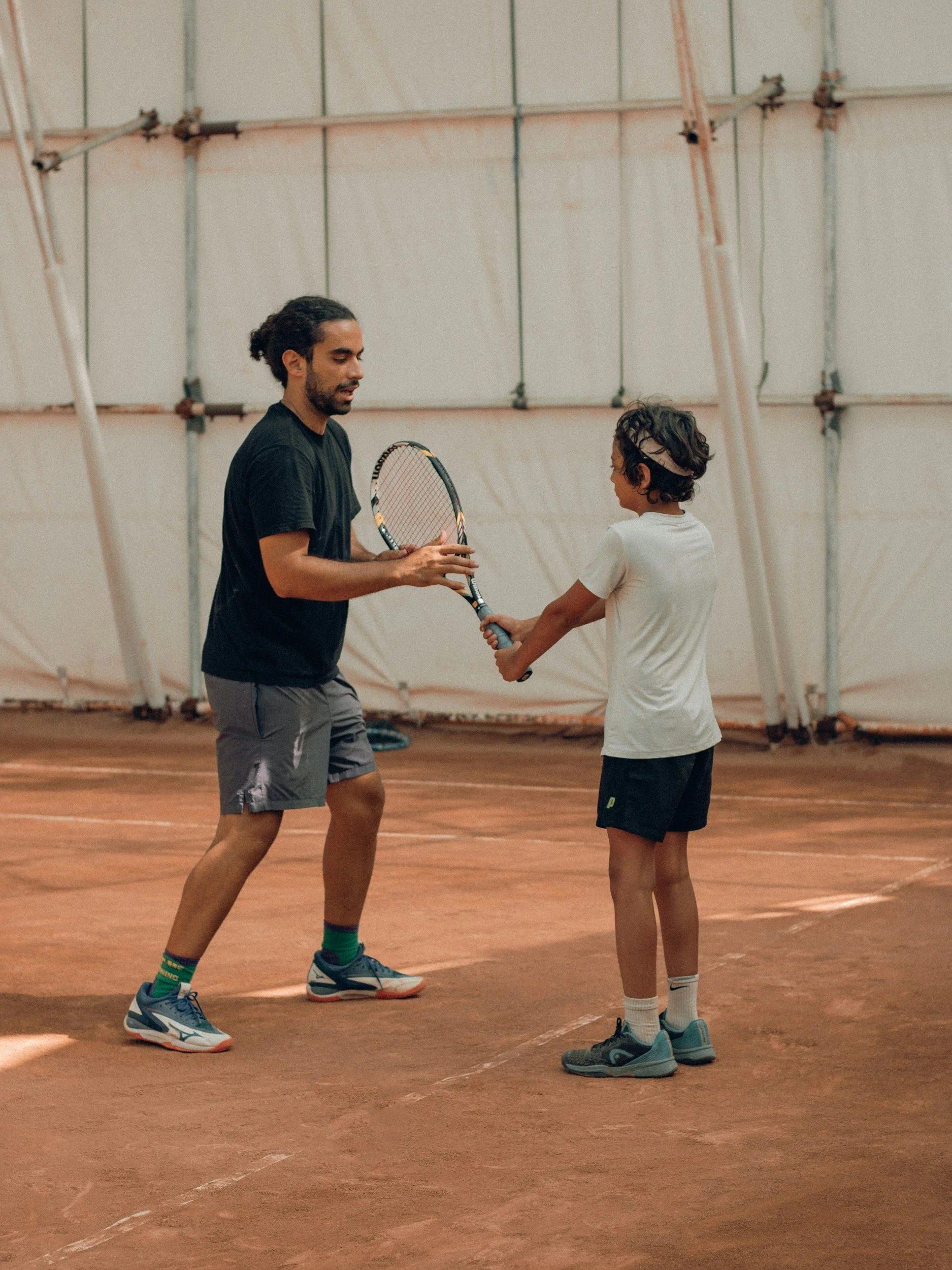 Two individuals playing tennis on an indoor clay court. The adult male is in black athletic attire, holding a tennis racket, and appears to be instructing the young boy, who is in a white T-shirt and black shorts, also holding a tennis racket.