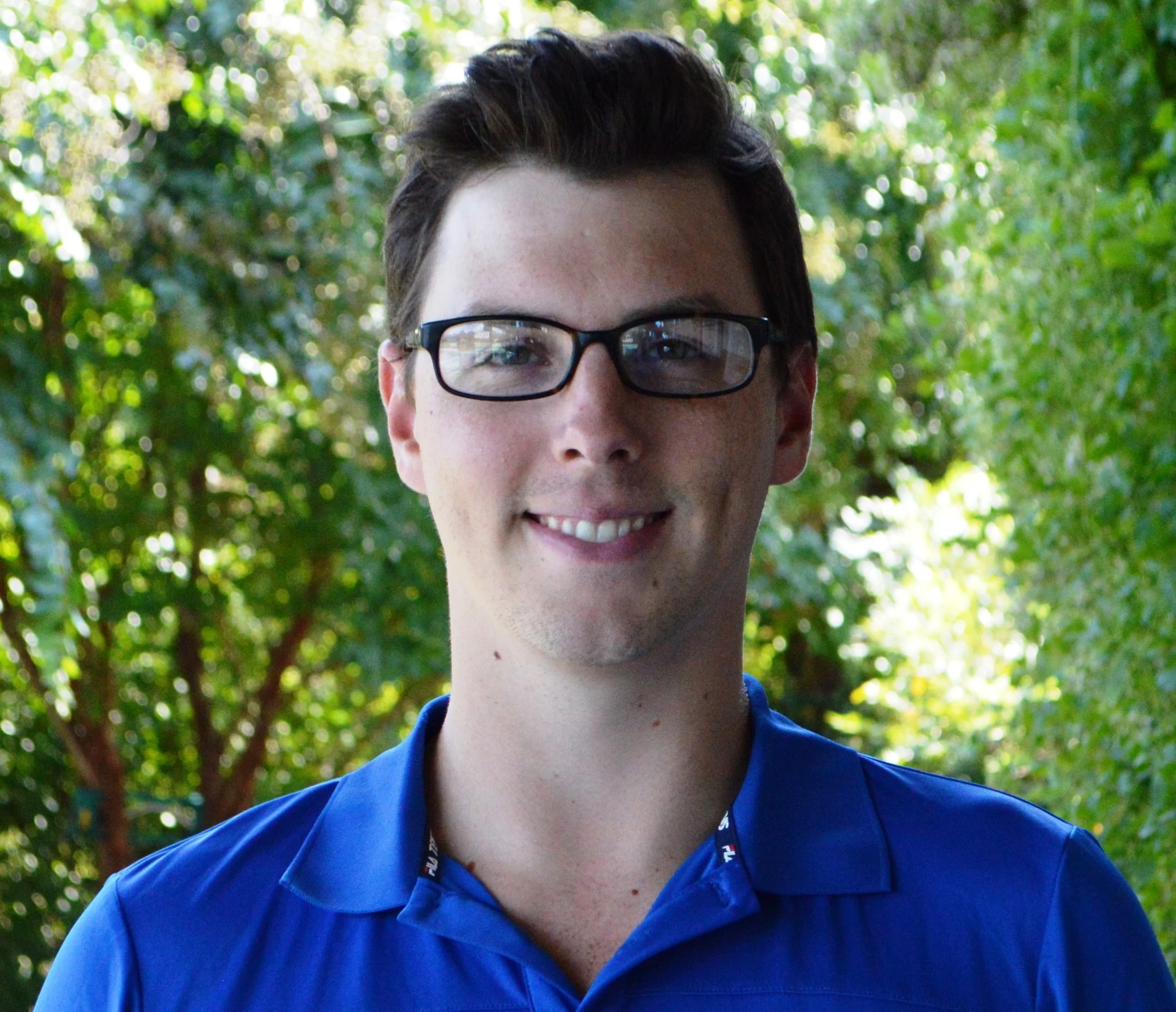 Close-up of a young man with short dark hair, wearing black-rimmed glasses, smiling outdoors among green trees, dressed in a blue collared shirt.