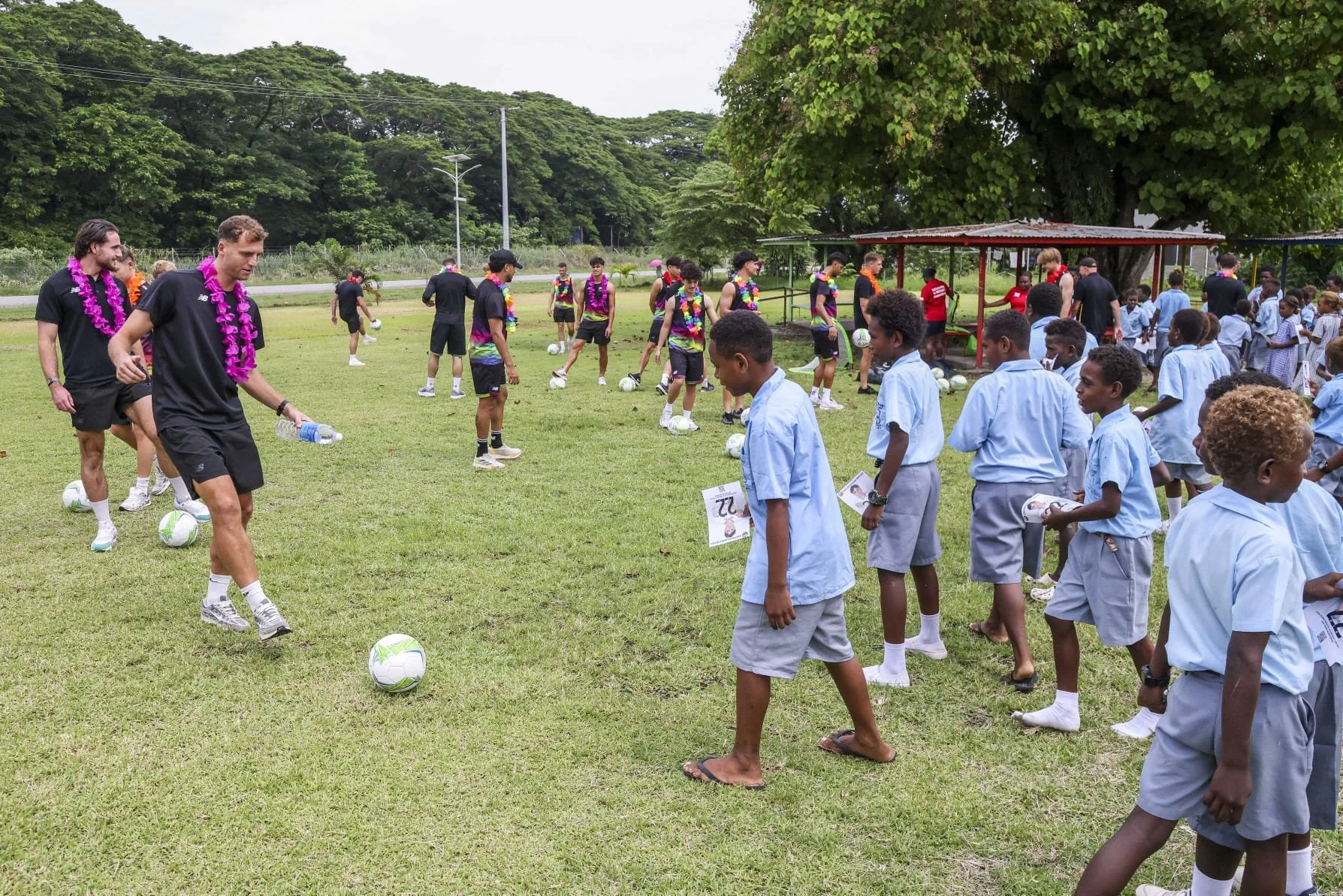 Children in school uniforms standing in line with soccer balls while adults in sportswear train on a grassy field under a tree, with other children and people in the background.