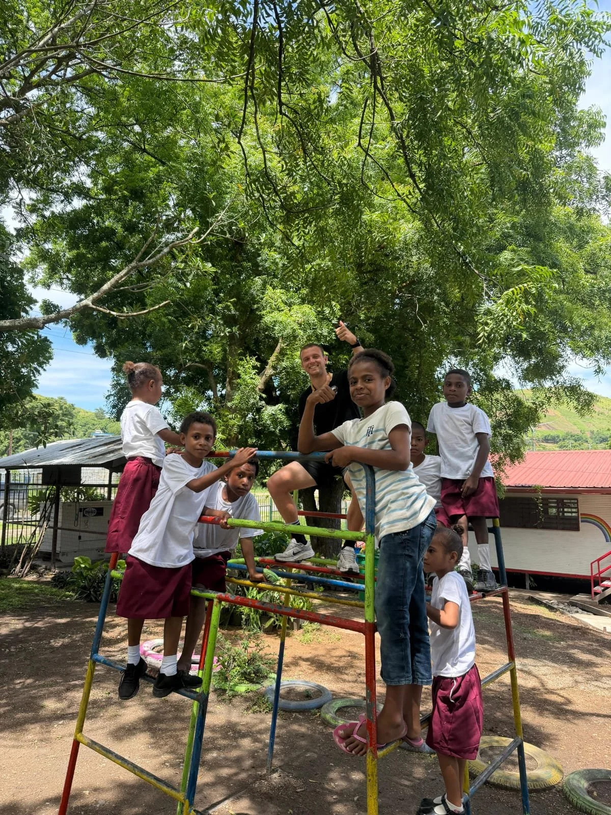Children and a man playing on a colorful jungle gym under a large tree outdoors. The children are wearing white shirts and maroon skirts or shorts, and the man is smiling and giving a thumbs-up.