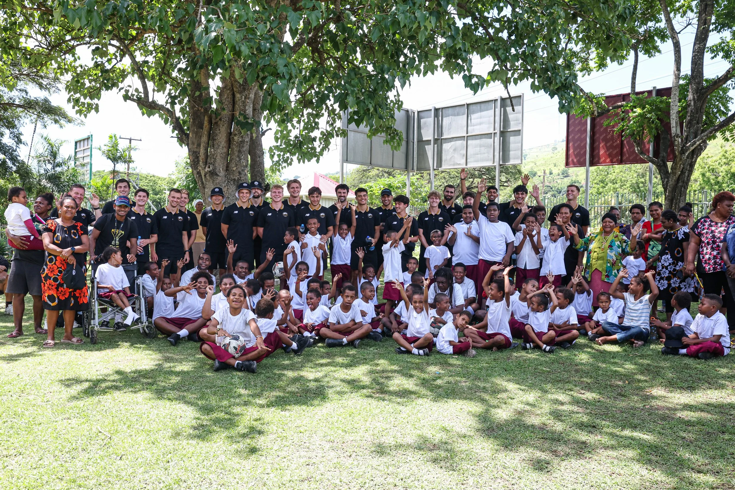 Whole team visit school and play football. Group photo after fun playing, gathered outdoors under a large tree, some children sitting on the grass and others standing.