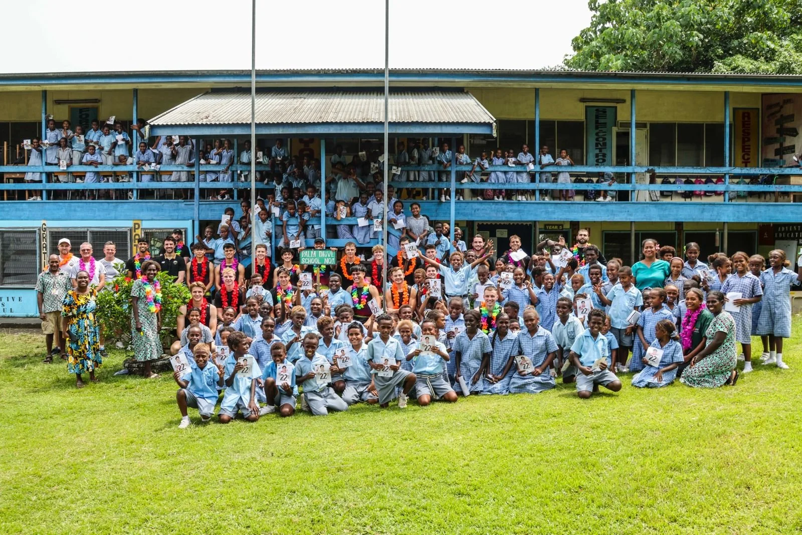 Large group of students, teachers, and visitors gathered outside a school building, celebrating a graduation or school event on a sunny day.