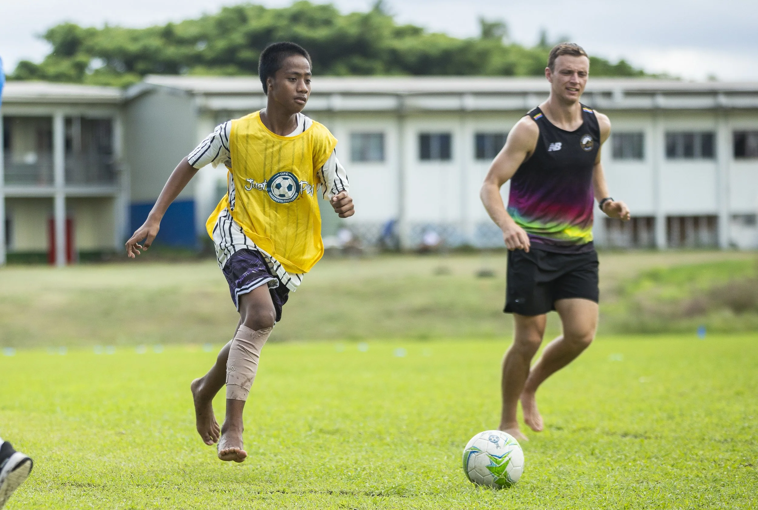 A young boy and a man running on a grassy soccer field with a white soccer ball at his feet. The boy is wearing a yellow vest and shorts, and the man is wearing a black and purple sleeveless shirt and black shorts.