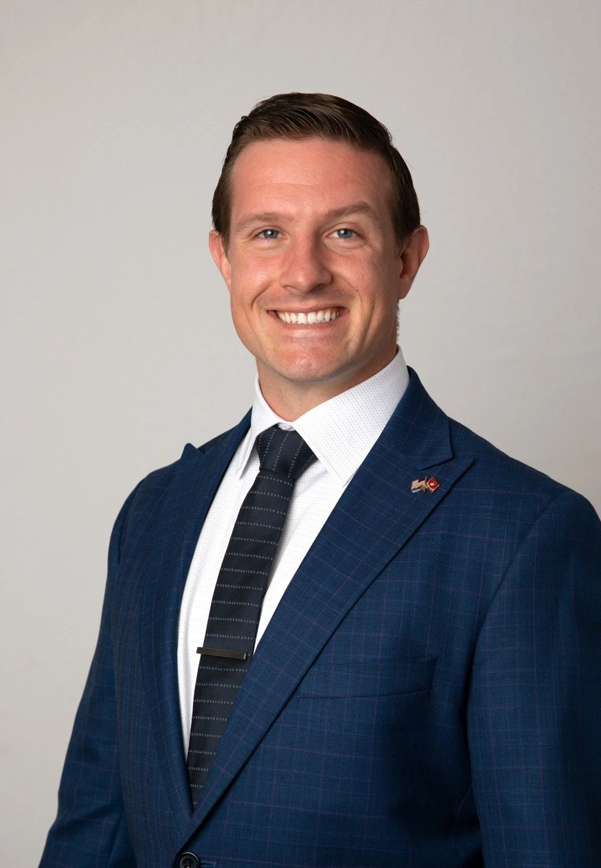 A smiling man in a blue plaid suit, white shirt, and striped tie, with an American flag pin on his lapel, posing against a light gray background.