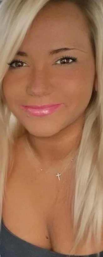 Close-up of a smiling, spiritual blonde woman with pink lip gloss, wearing a silver cross necklace.