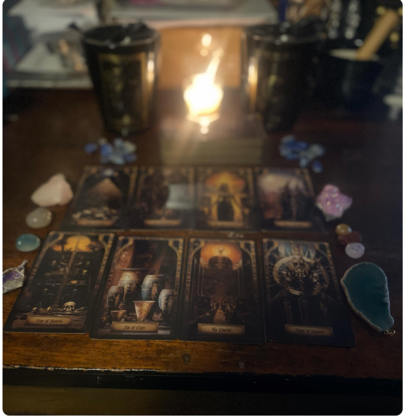 A tarot card reading setup with eight tarot cards laid out on a wooden surface, surrounded by various crystals and stones. A candle provides soft lighting, creating a mystical atmosphere.