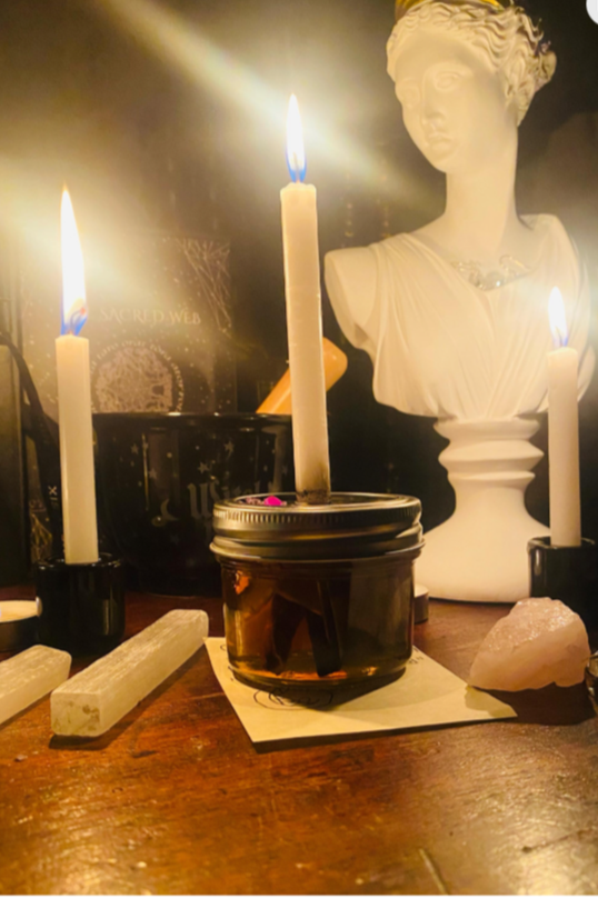 A spiritual or ritual setup with three lit candles, a jar candle, a white bust sculpture, and various stones on a wooden surface.