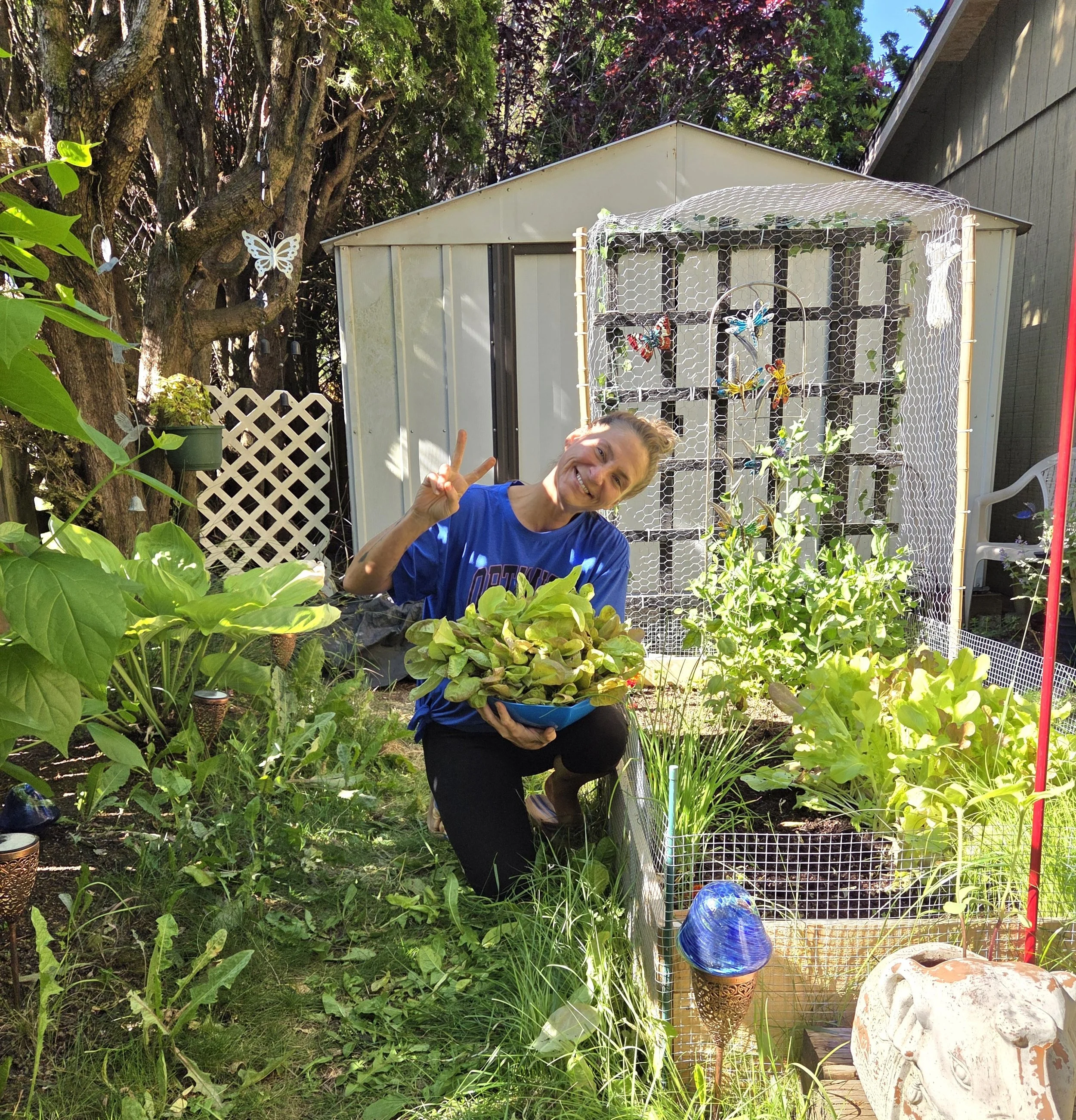 A woman smiling and making a peace sign in a backyard garden. The garden has lush green plants, trees, and decorative butterflies on a fence. There is a small shed and some garden ornaments, including a large pottery piece and a stack of blue glass bowls, in the background.