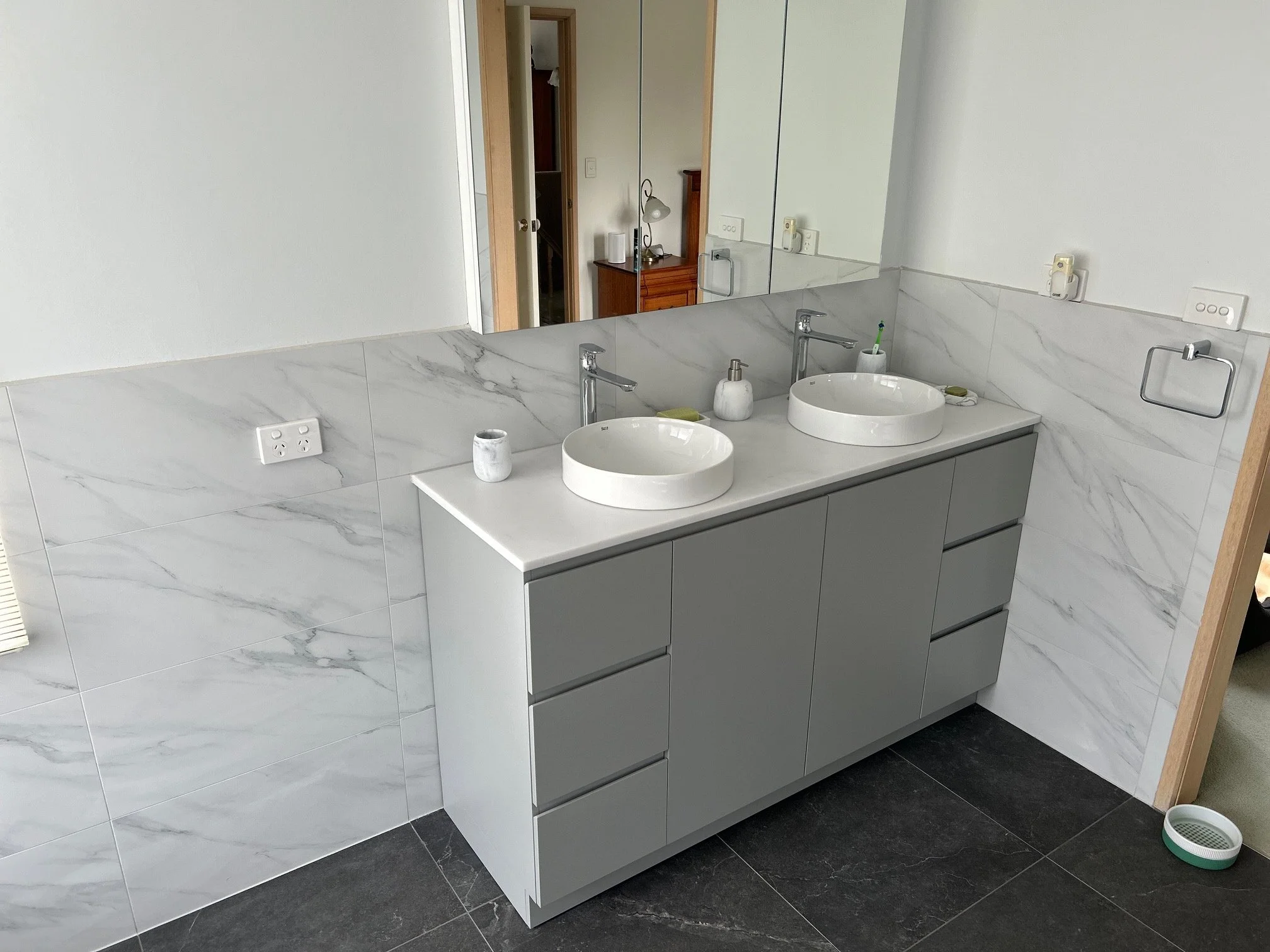 Modern bathroom vanity with two white vessel sinks, gray cabinets, marble-look tile walls, and mirrored backsplash